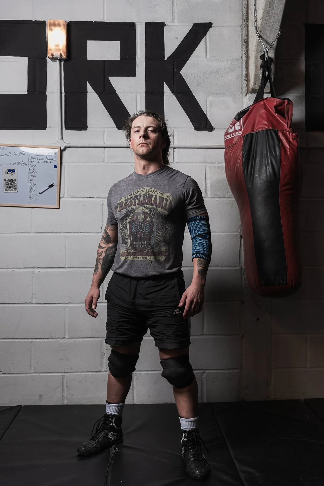 A wrestler in athletic shorts and a T-shirt standing in a gym. He is wearing knee pads and wrestling shoes, with a serious expression. Behind him is a punching bag hanging on the wall.