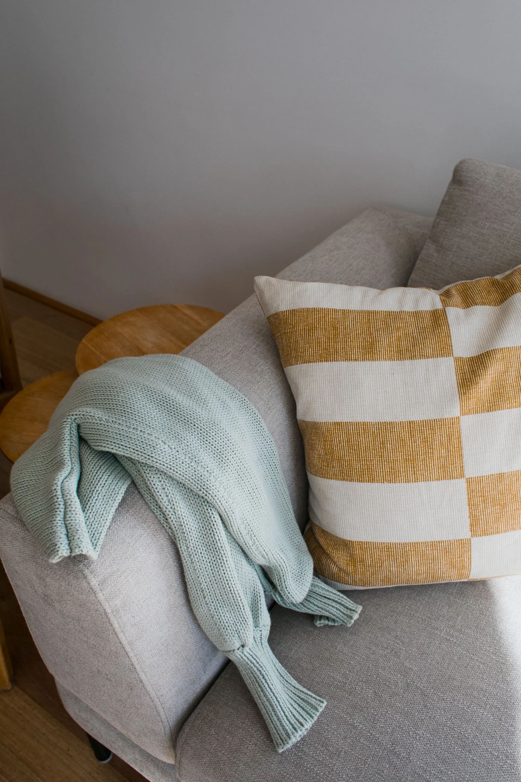 Close-up of a cozy corner with a beige upholstered armchair, a white and yellow plaid pillow, a mint green knitted blanket, a small wooden side table, and a plain gray wall background.