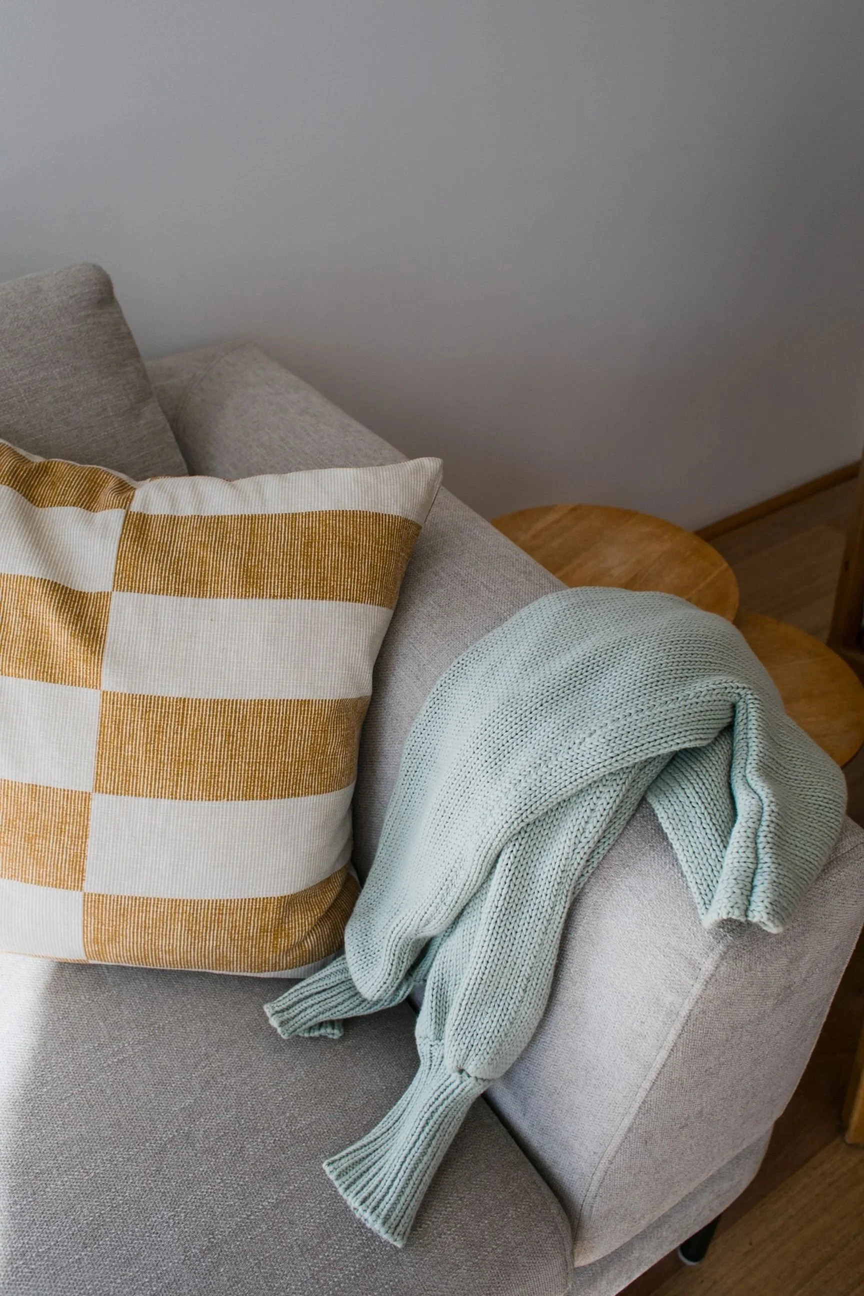 A beige couch with a yellow and white checkered pillow and a mint green knit blanket draped over the armrest. There is a small wooden round side table behind the couch.