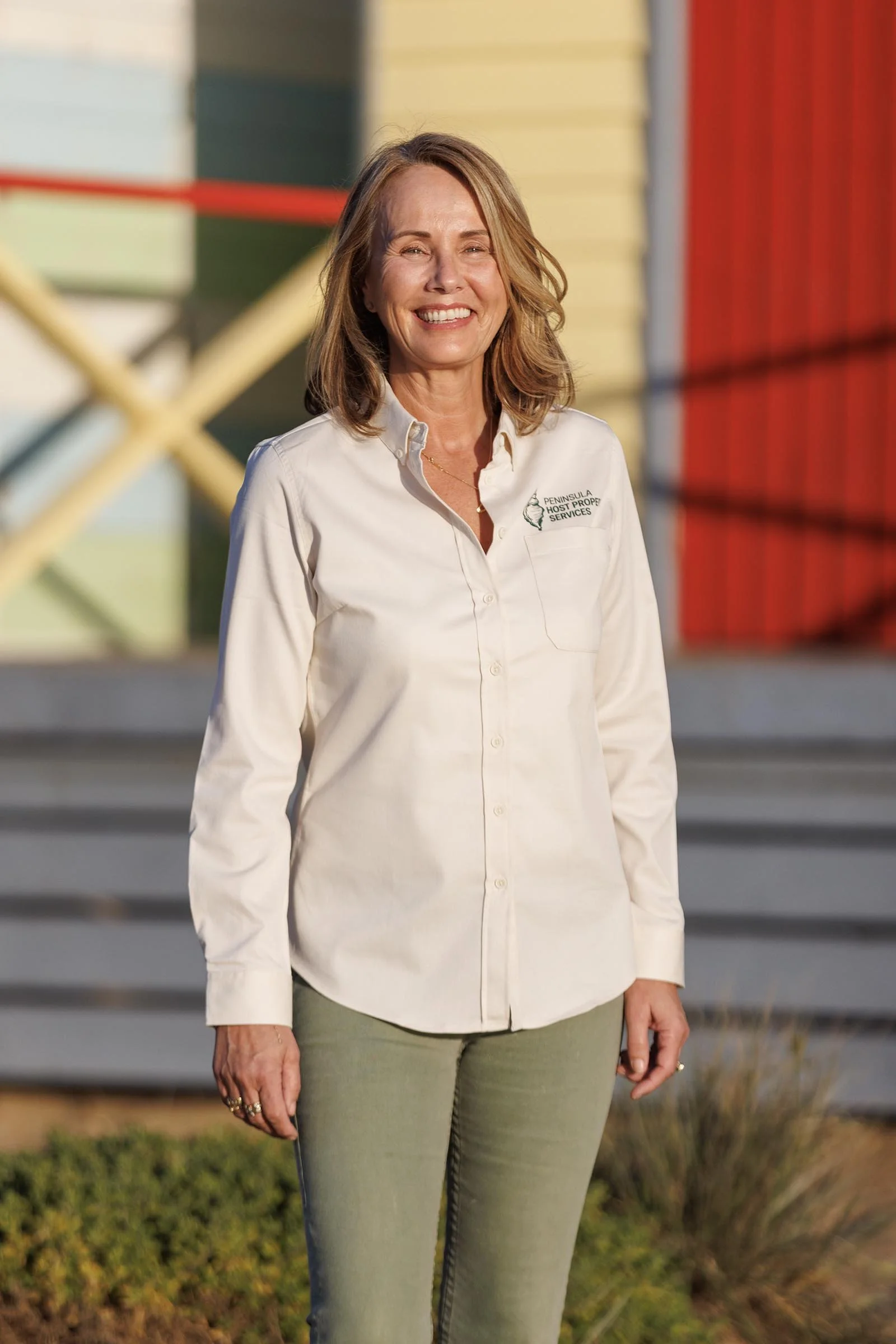 A woman with shoulder-length light brown hair, smiling, and wearing a white button-up shirt with a company logo, stands outdoors near a staircase with colorful walls in the background.