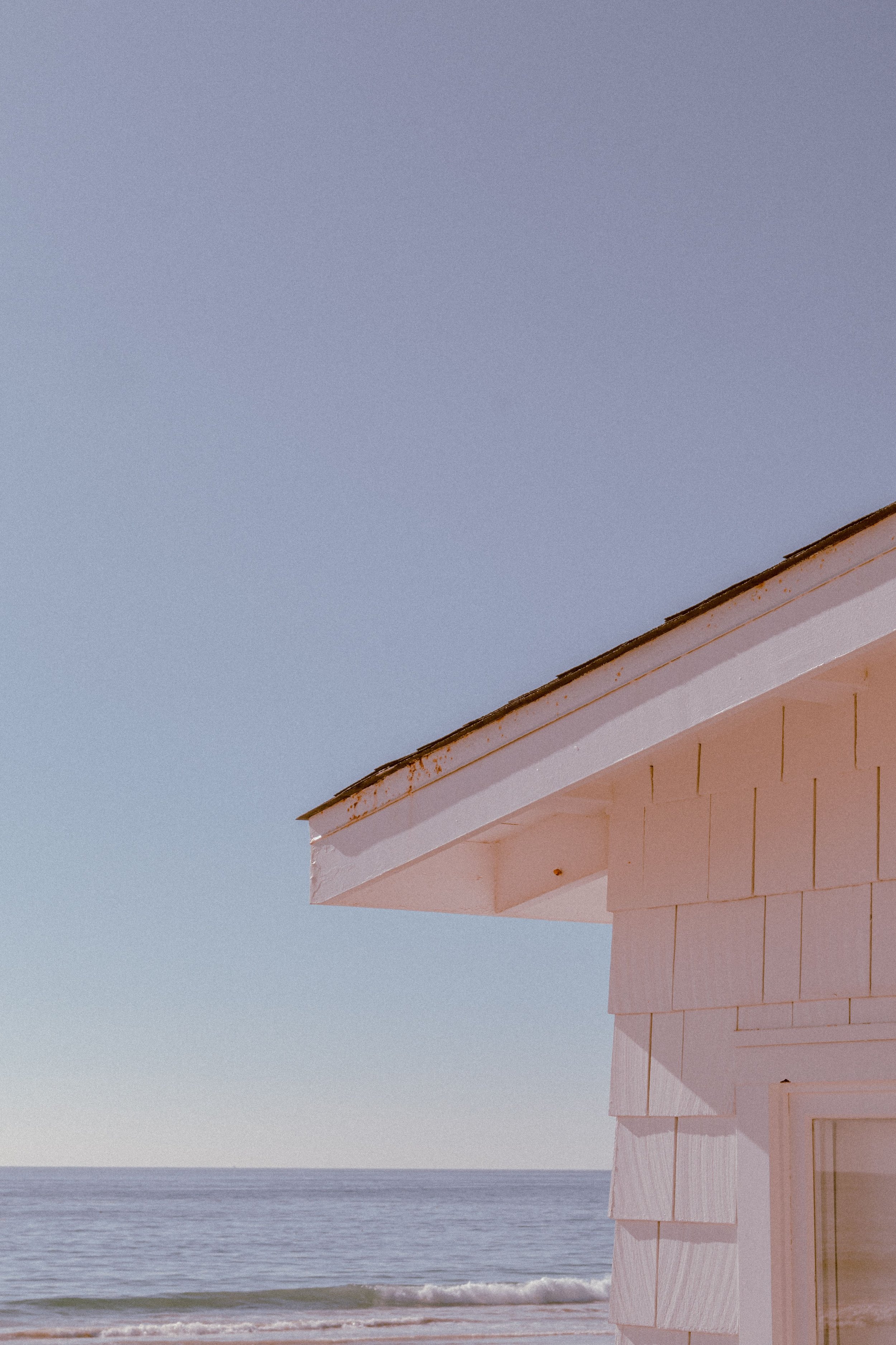 A house with white shingle siding and a rusted metal roof, facing the ocean during sunset or sunrise.