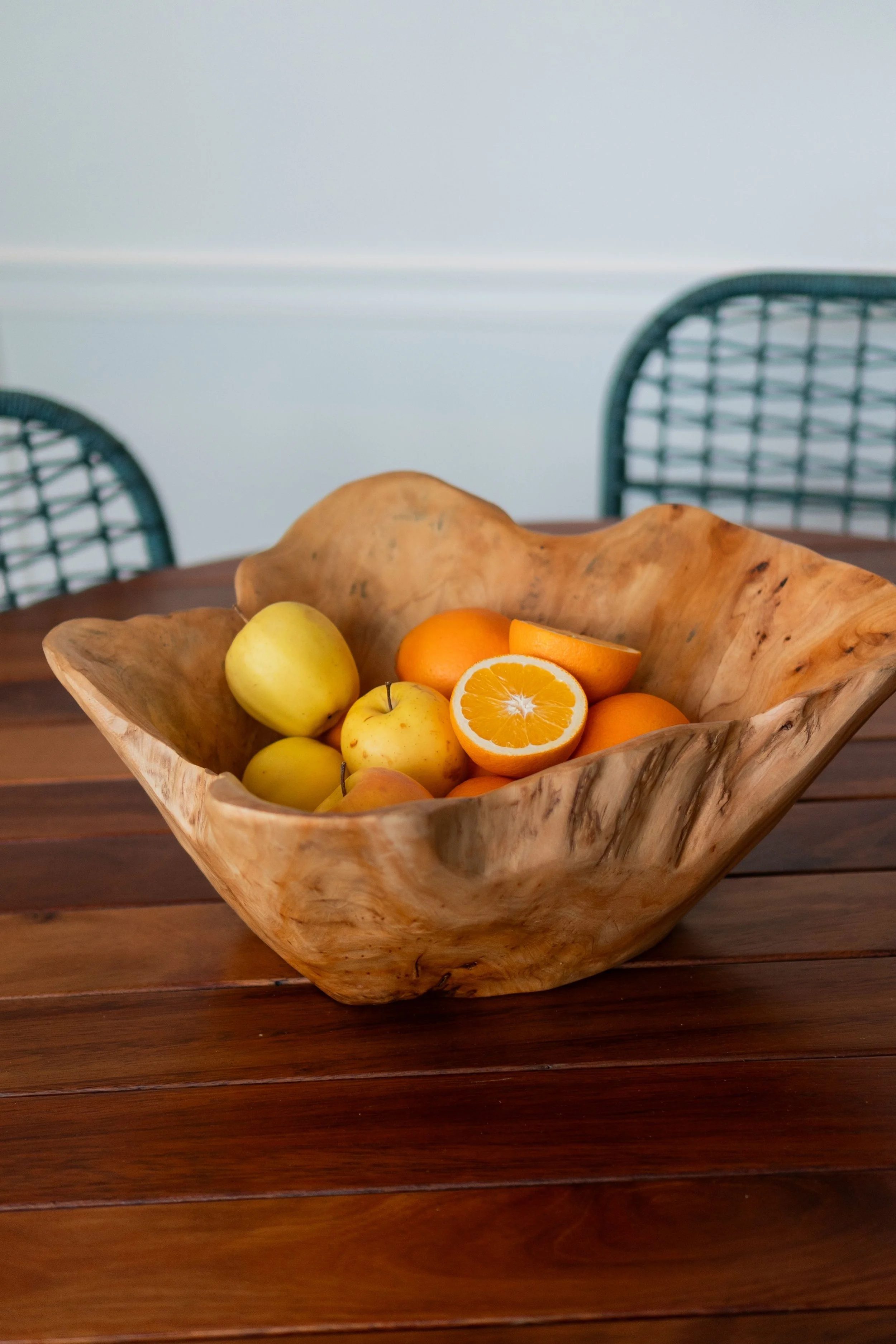 A wooden bowl containing yellow apples, oranges, and sliced oranges on a wooden table, with chairs in the background.