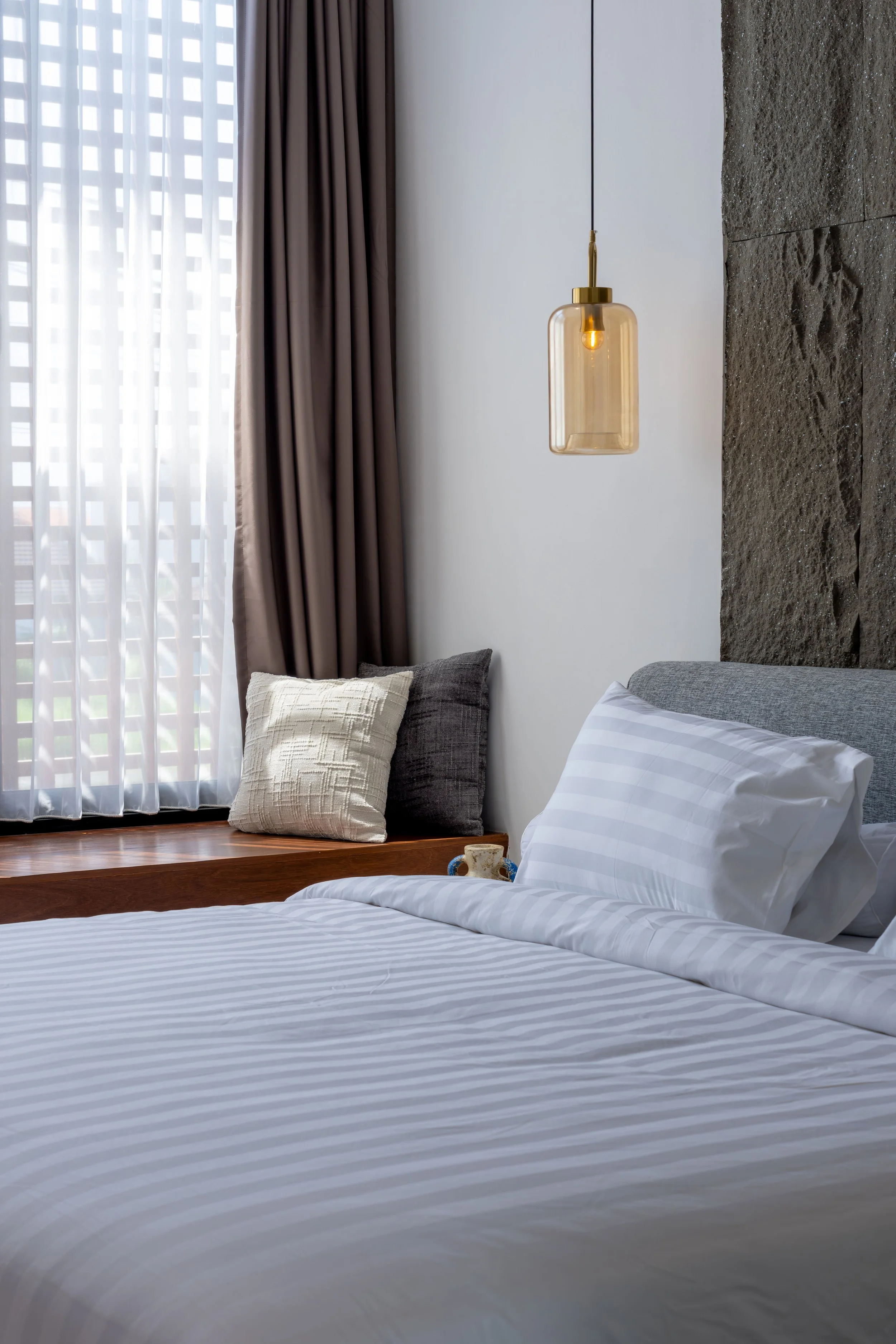 A neatly made bed with white striped sheets and pillows, next to a window with sheer curtains and brown drapes, a hanging glass light fixture, and a textured stone wall.