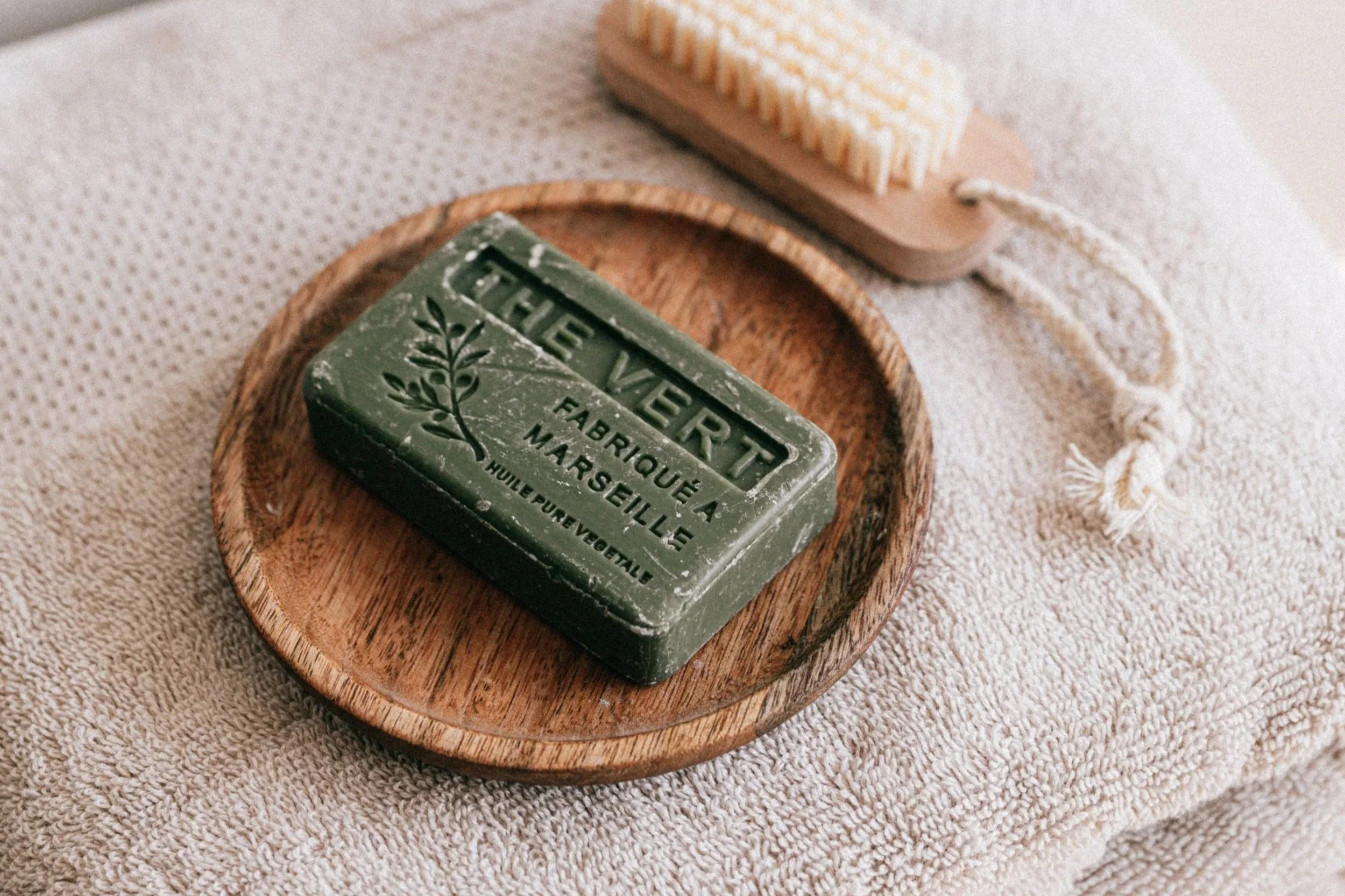 A green bar of soap with various embossed text, placed on a small round wooden tray, with a beige textured fabric underneath.