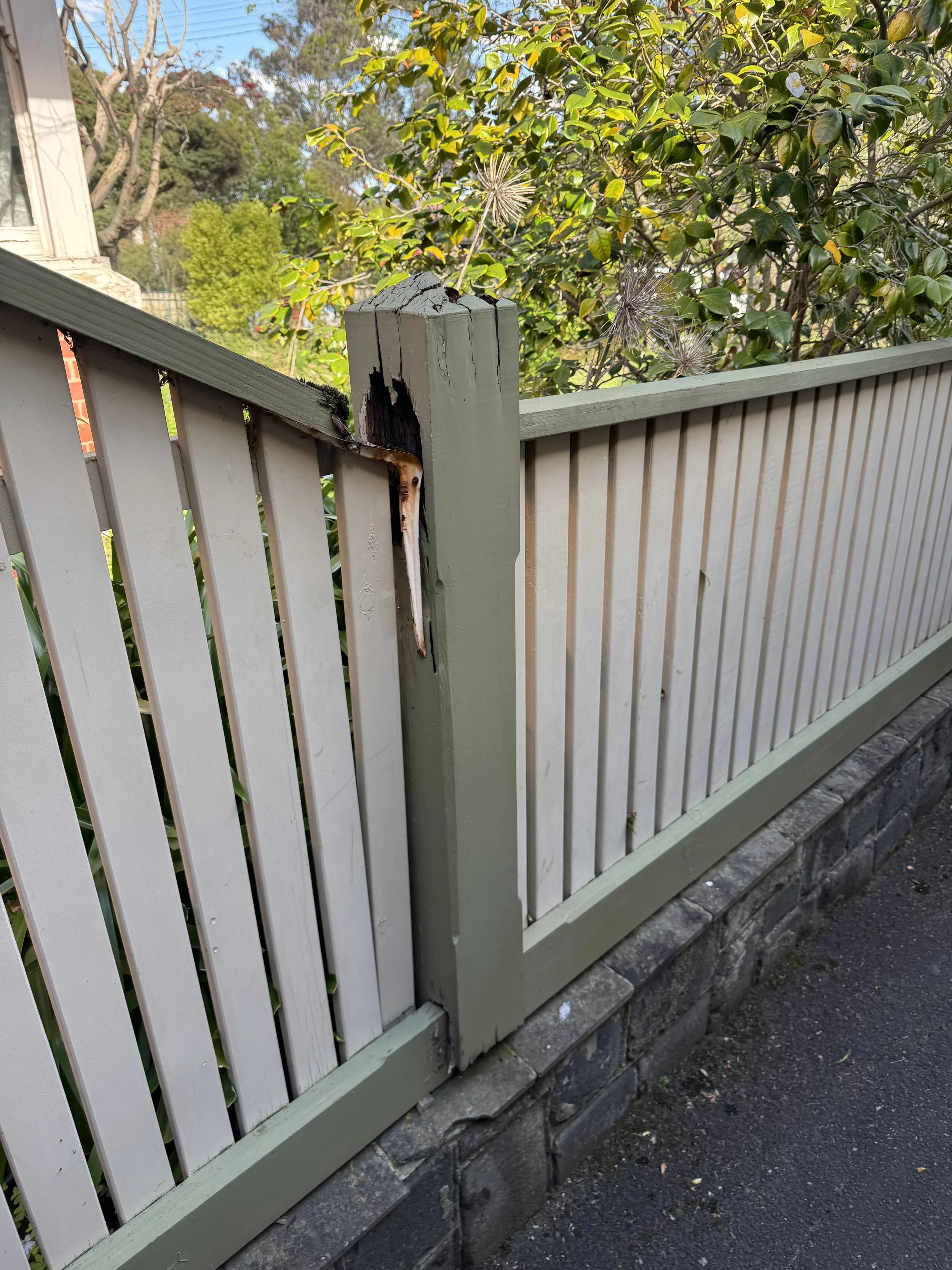 Wooden white picket fence with a damaged corner, adjacent to a stone wall, on a sidewalk with greenery and trees in the background.