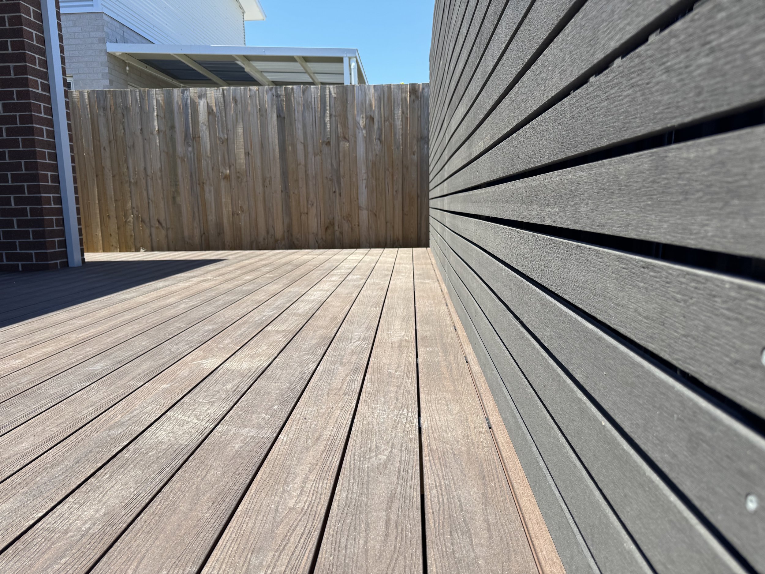 View of a wooden deck with a wooden privacy fence, a section of house siding, and part of a residential area with a small roof extension and bright blue sky.