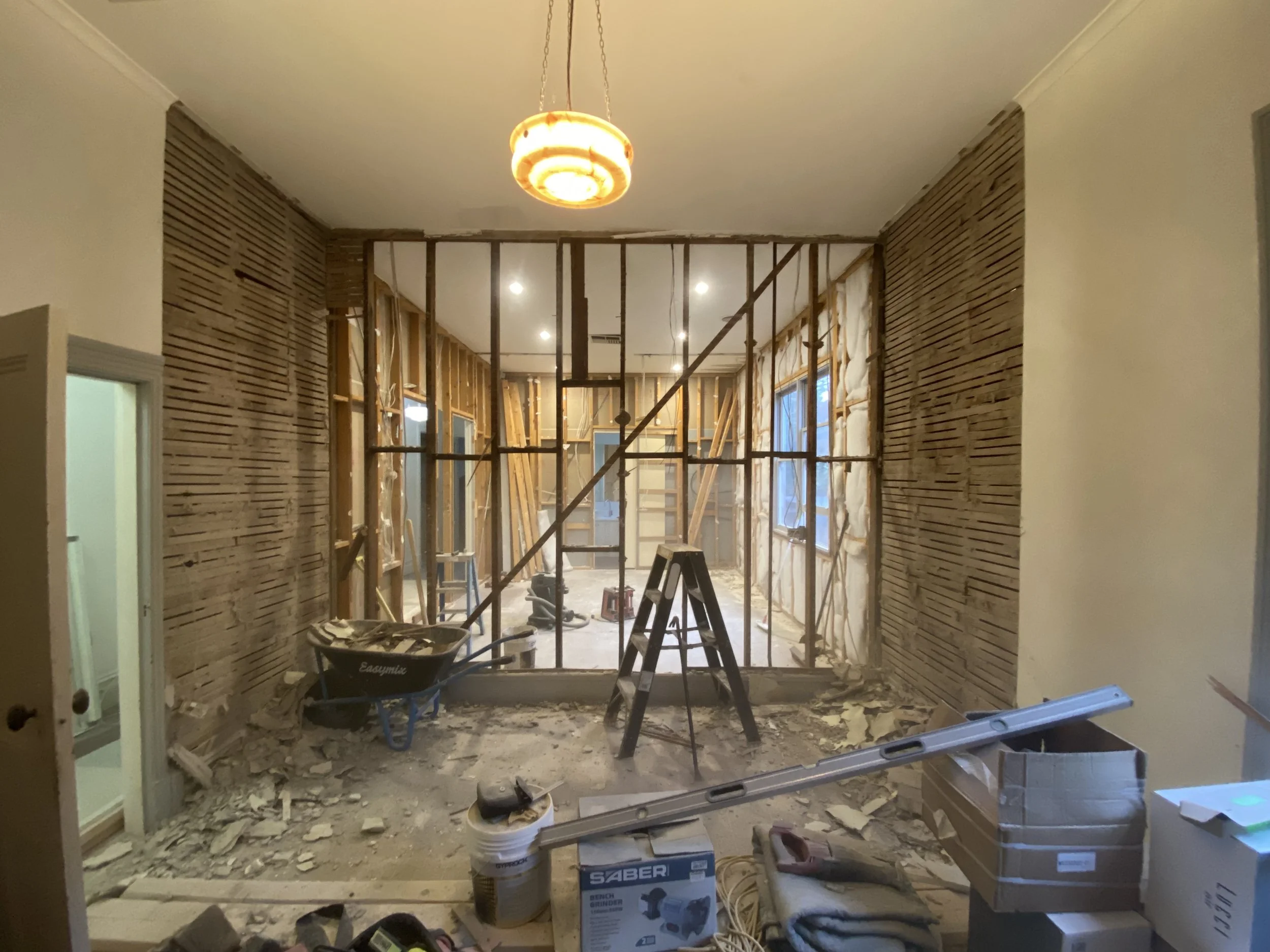 Room under renovation with wooden framing and construction tools, debris on the floor, and a chandelier hanging from the ceiling.