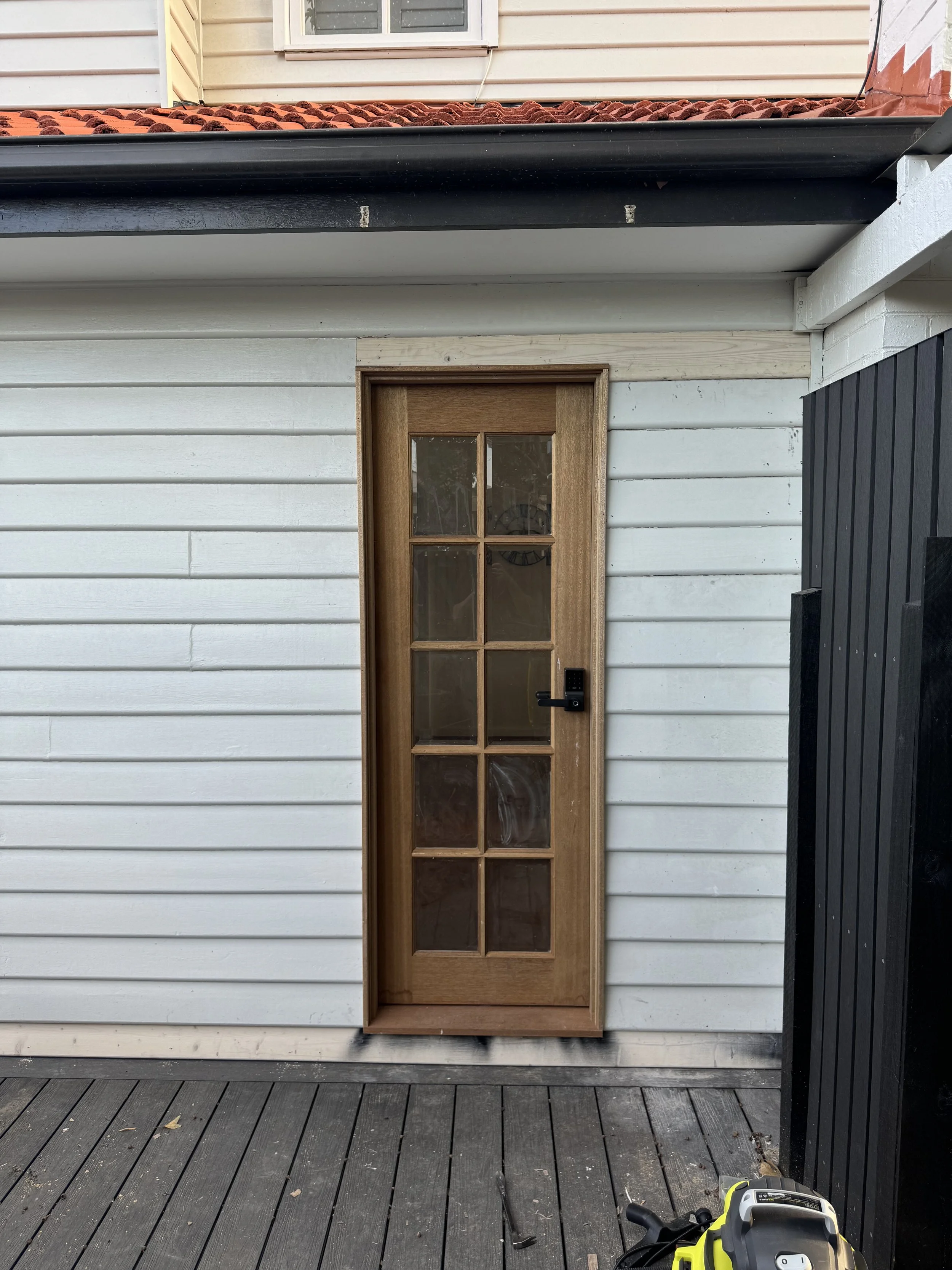 The image shows a small wooden door with glass panels, set into a white wall with horizontal siding. There is a black and yellow power tool on the dark wooden deck floor in the foreground.