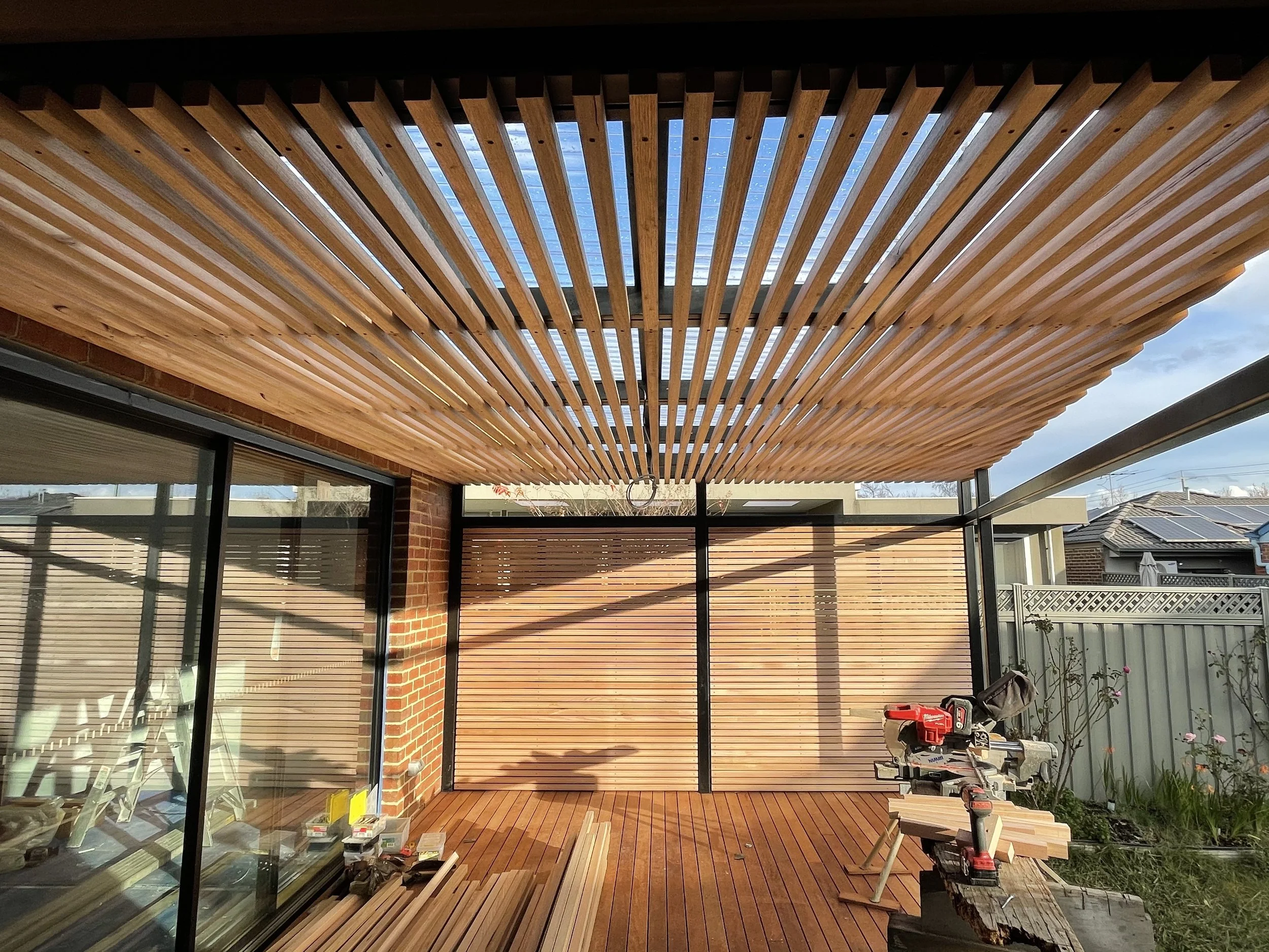 Construction of a wooden patio cover or pergola attached to a house, with tools and materials nearby, and a partly cloudy sky in the background.