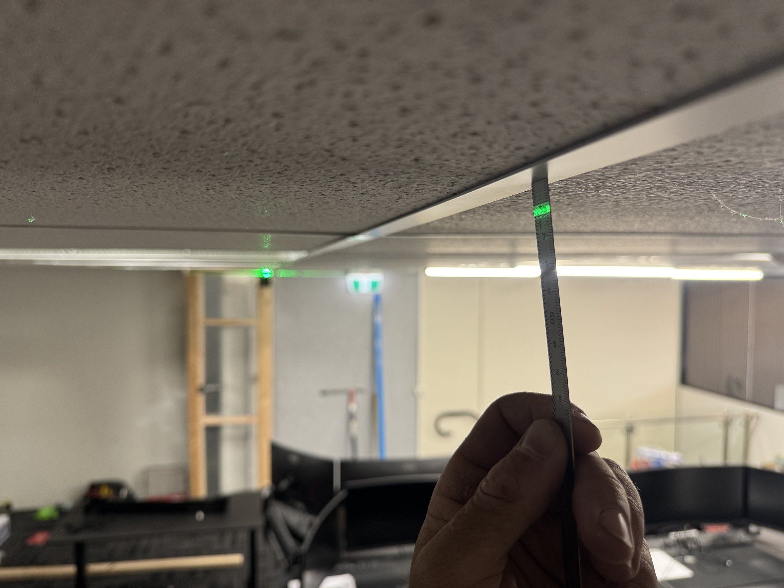 Person holding a measurement ruler under a textured ceiling with multiple green laser points projecting onto it, in an indoor setting with shelves and fluorescent lights.