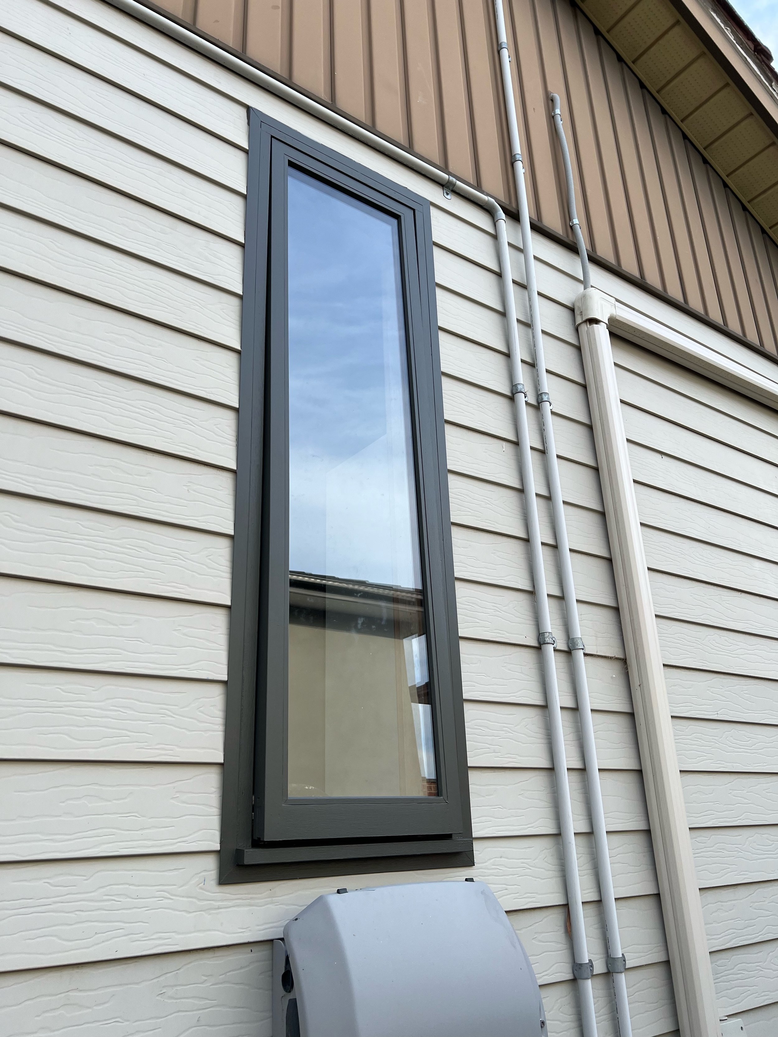 Exterior of a house with beige and brown siding, a rectangular window with a dark gray frame, and piping running vertically along the wall.