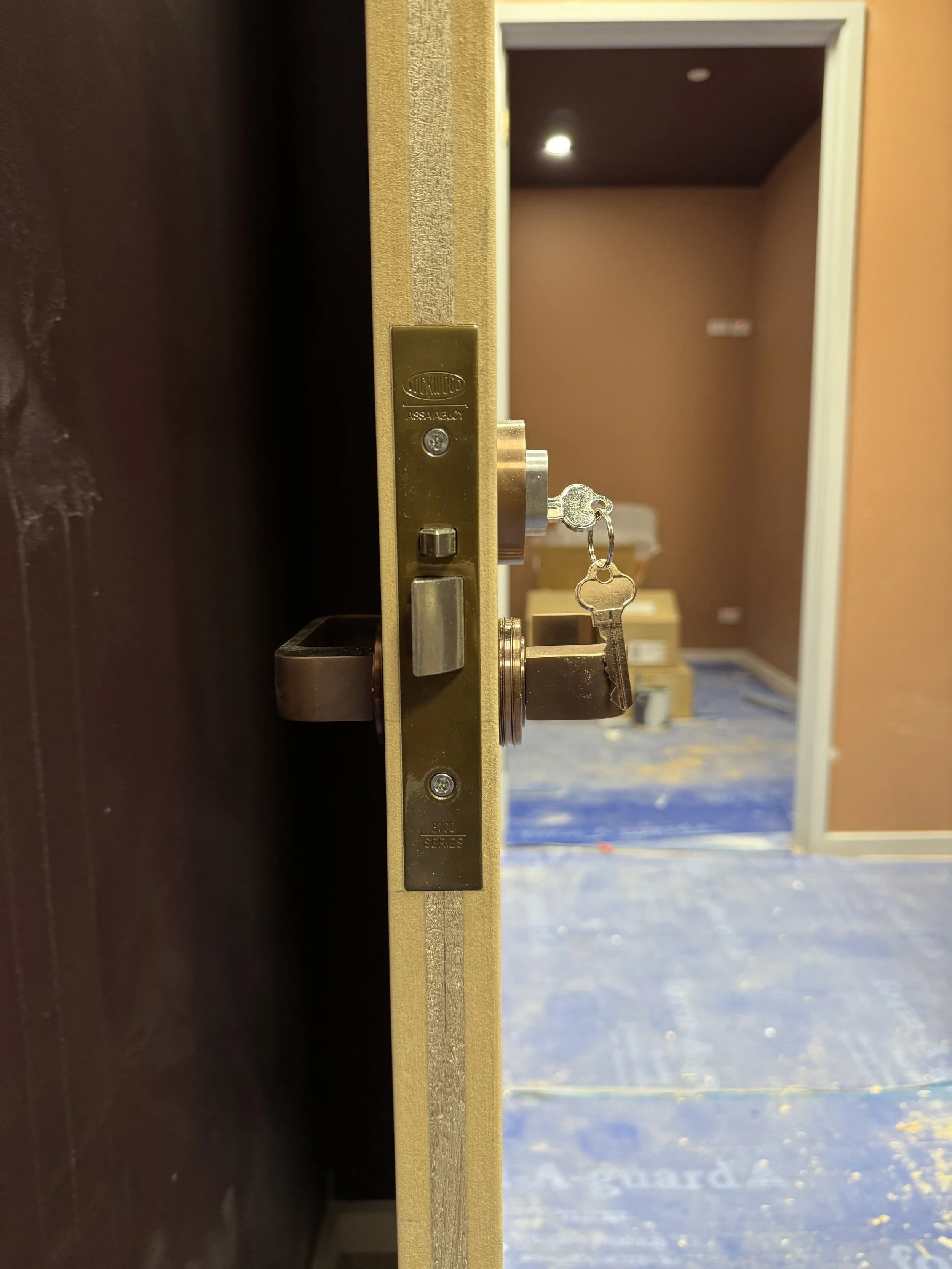 Close-up of a door lock with a key hanging from the lock, revealing a background hallway with a chair and blue carpet.