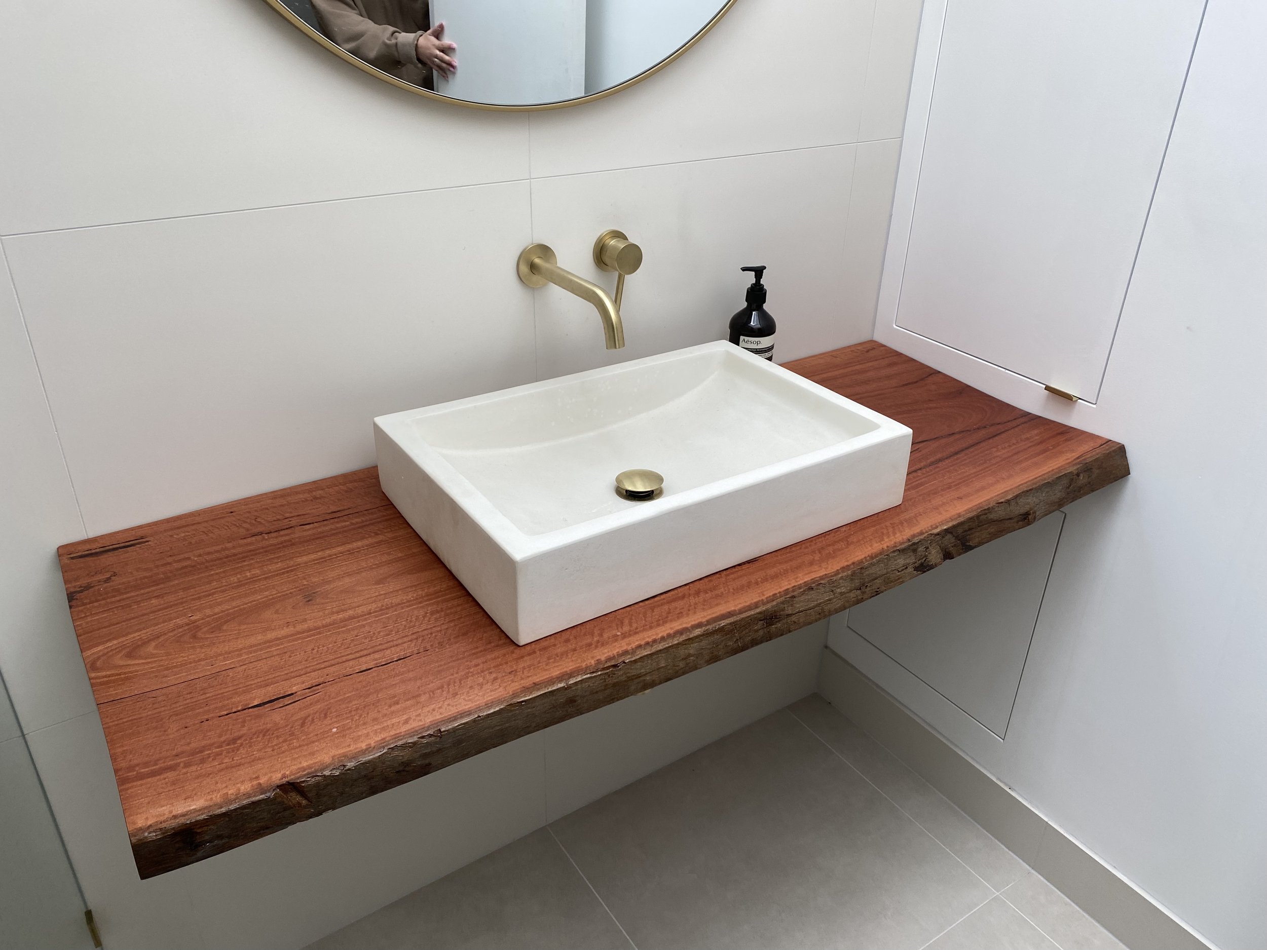 Modern bathroom with a white rectangular vessel sink on a wooden countertop, a gold wall-mounted faucet, a black soap dispenser, a mirror, and white tile walls.