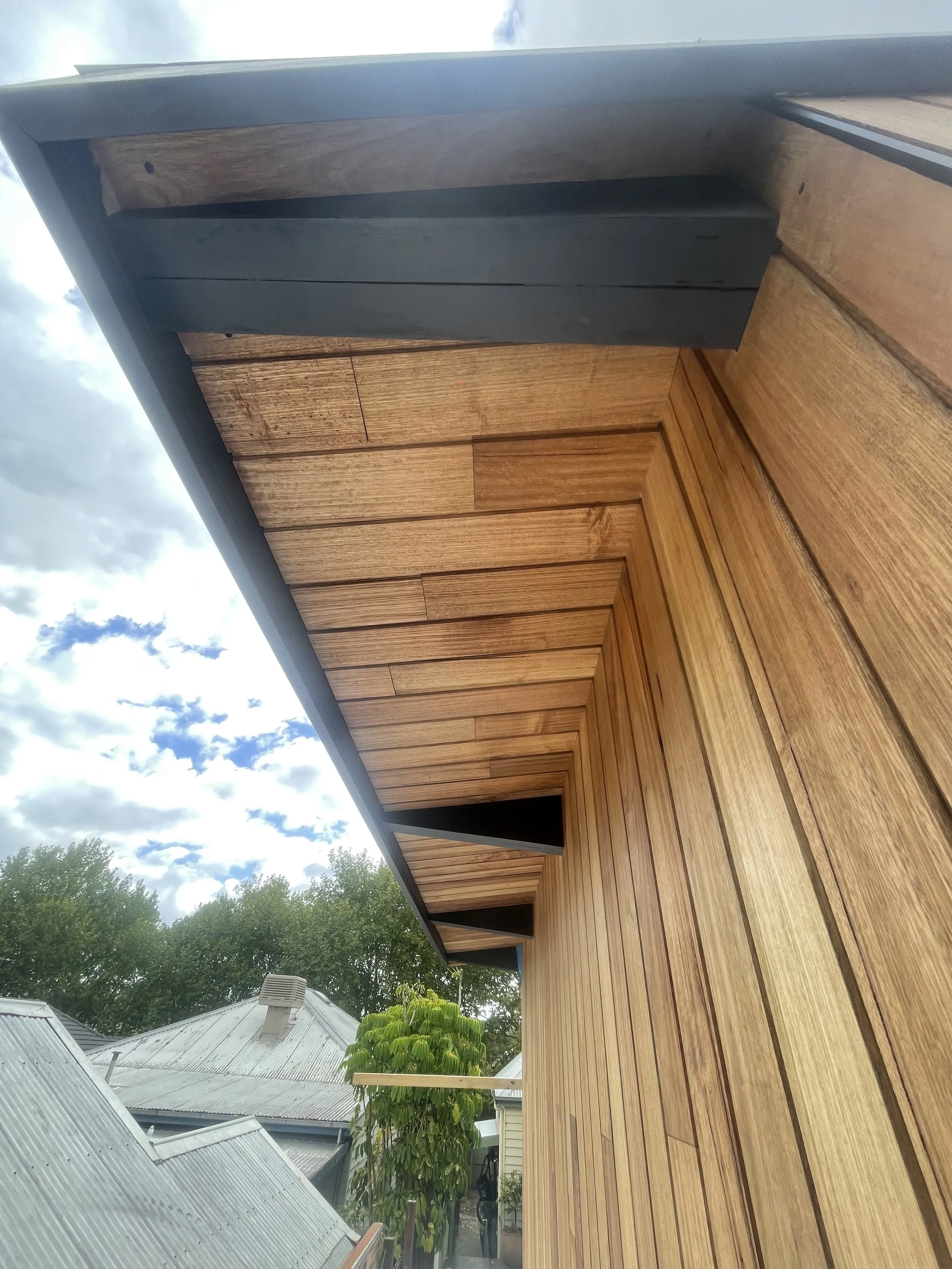 View of a wooden deck and house exterior with two small openings in the wall, surrounded by trees and other rooftops, under a partly cloudy sky.