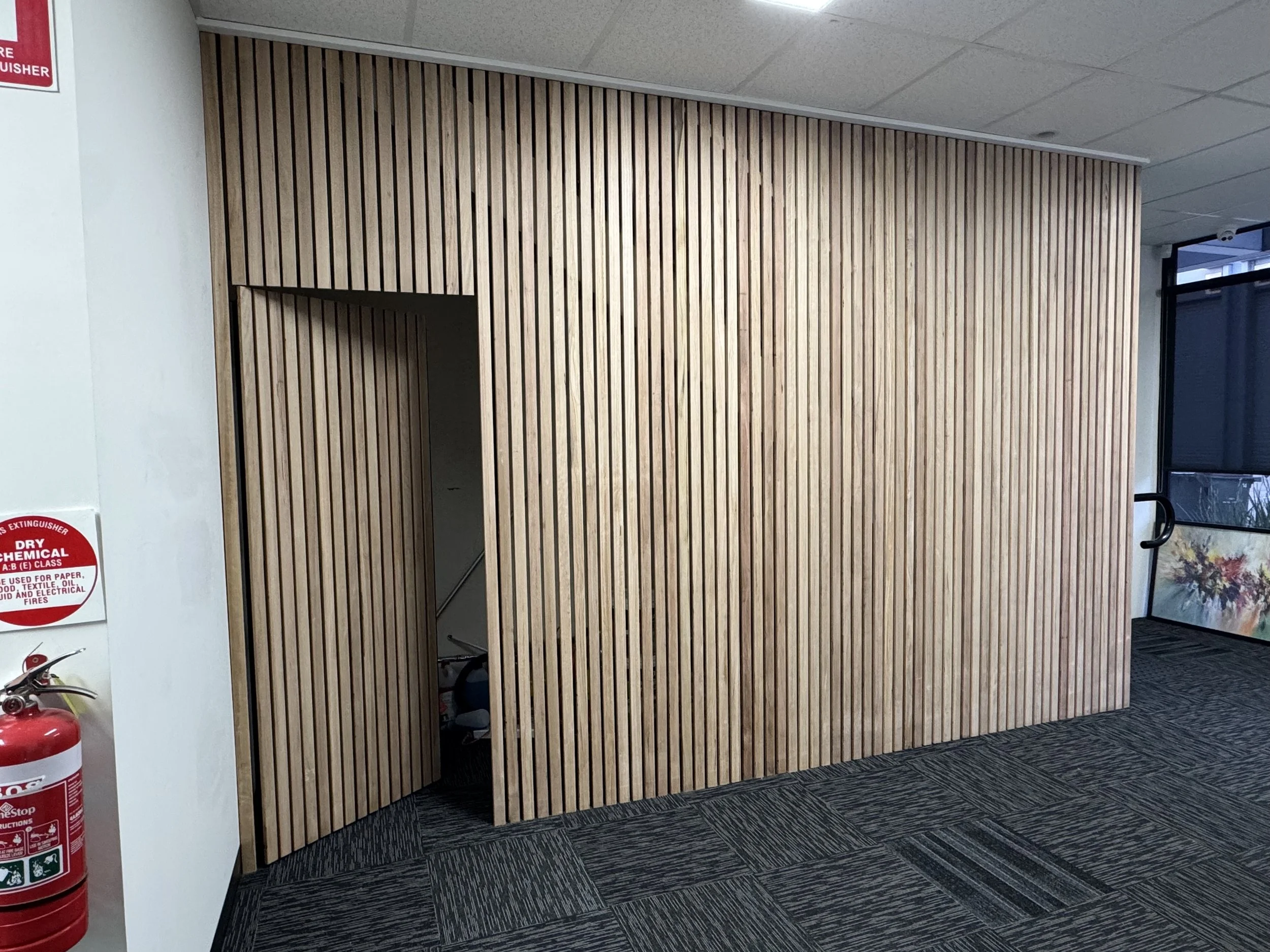 Wooden slat partition wall in an indoor space, with a doorway cutout, located near a fire extinguisher and a window.