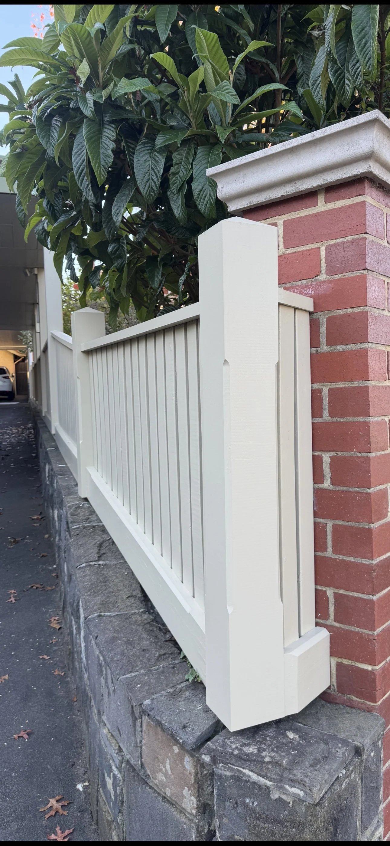 Close-up of a white wooden fence with vertical slats, supported by a brick pillar with a concrete cap, alongside a sidewalk with fallen leaves and a large leafy shrub in the background.