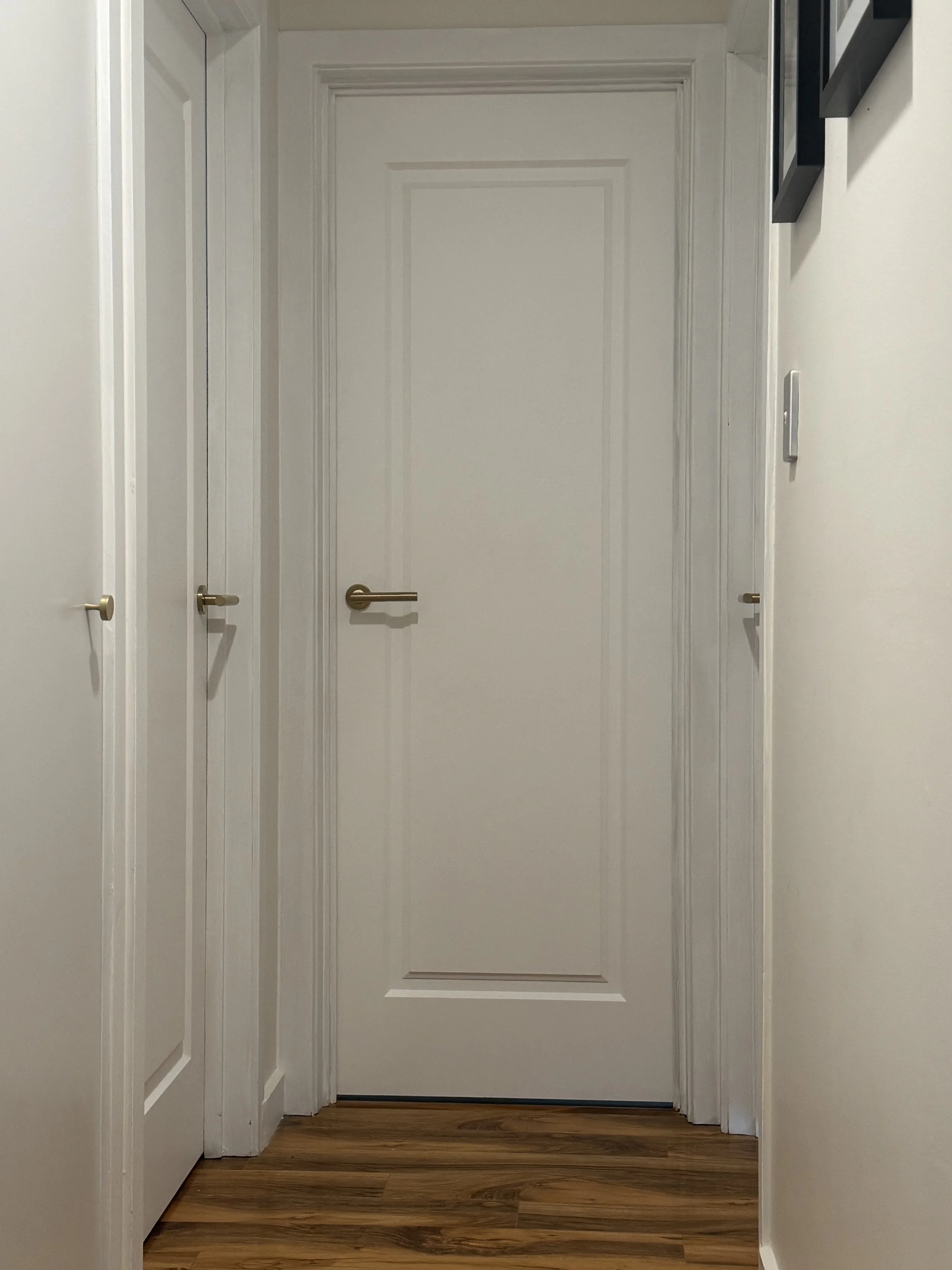 A hallway with a closed white door at the end, flanked by white walls and two additional doors with brass handles on either side. The floor is wood.