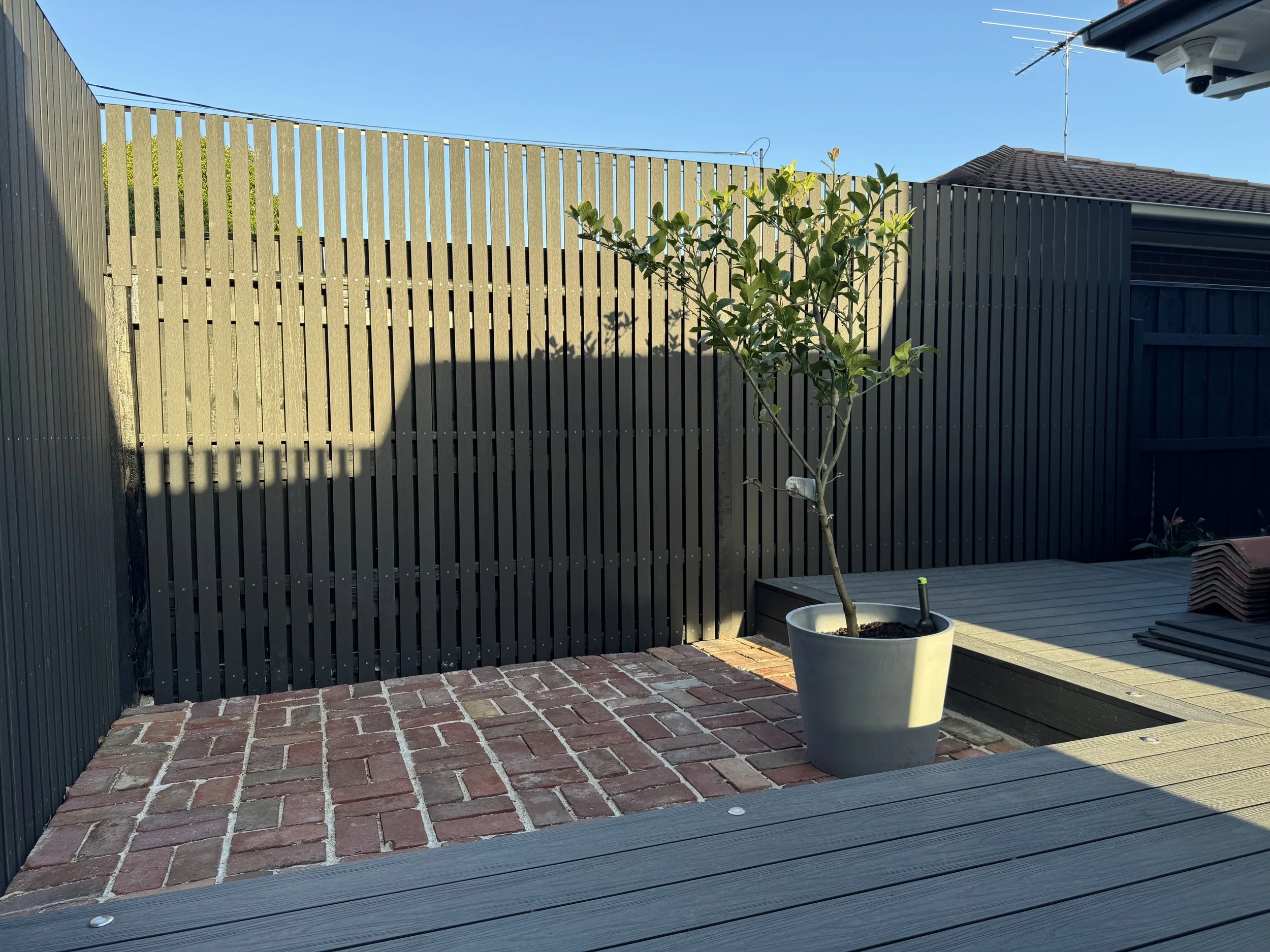 A backyard patio with red brick paving, a potted tree in the center, a wooden deck, a dark gray wooden fence, a house with a roof and satellite dish, and a blue sky.