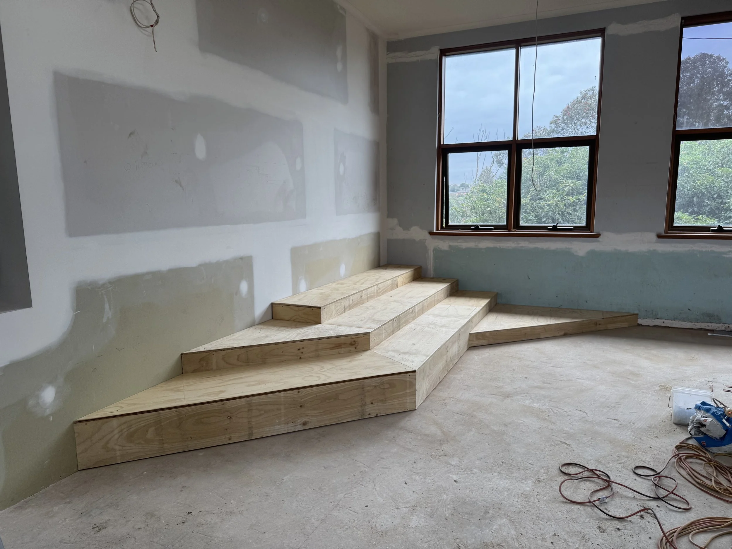 Interior of a room under construction with new wooden stairs and large windows showing trees outside.