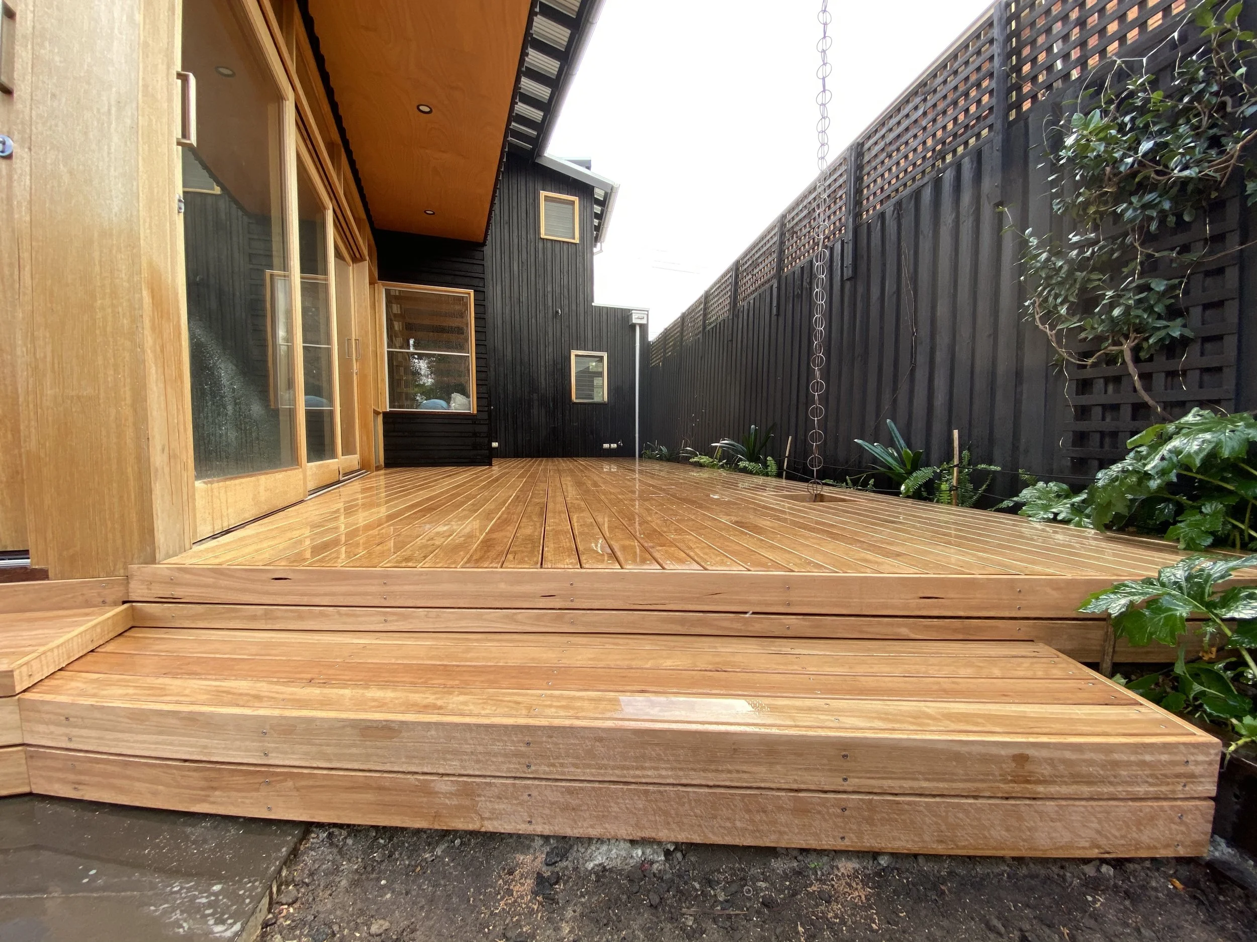 A newly built wooden deck at the back of a house, with a black wooden fence, plants on the right, and a house with wooden and black siding with windows on the left.