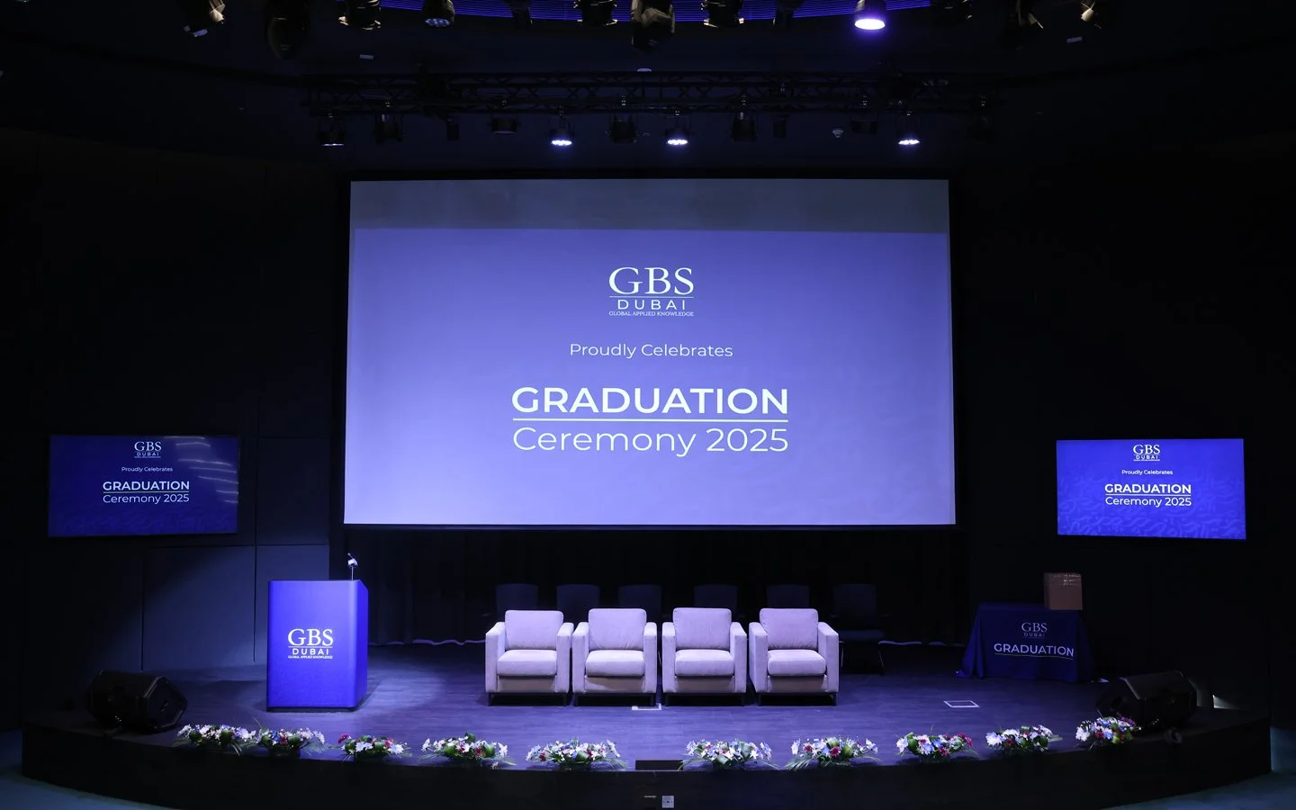 Stage set for the GBS Dubai graduation ceremony 2025, with a large central screen displaying the event details, two smaller screens on either side, a podium labeled GBS Dubai, six chairs arranged in a row, a table with a GBS Dubai GRAUDATION cloth, and floral decorations along the front of the stage.