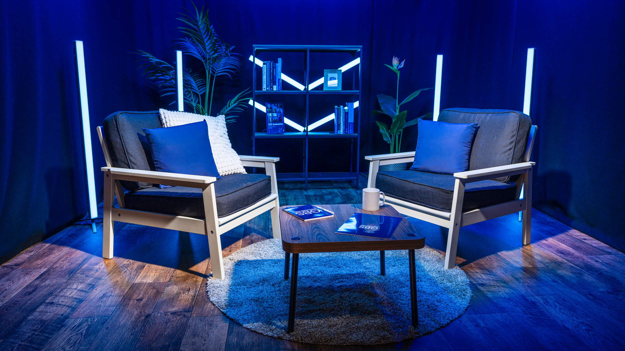 Two black armchairs with white frames and dark cushions, placed around a small wooden coffee table with magazines and a white mug, set in a blue-lit room with plants and bookshelves.