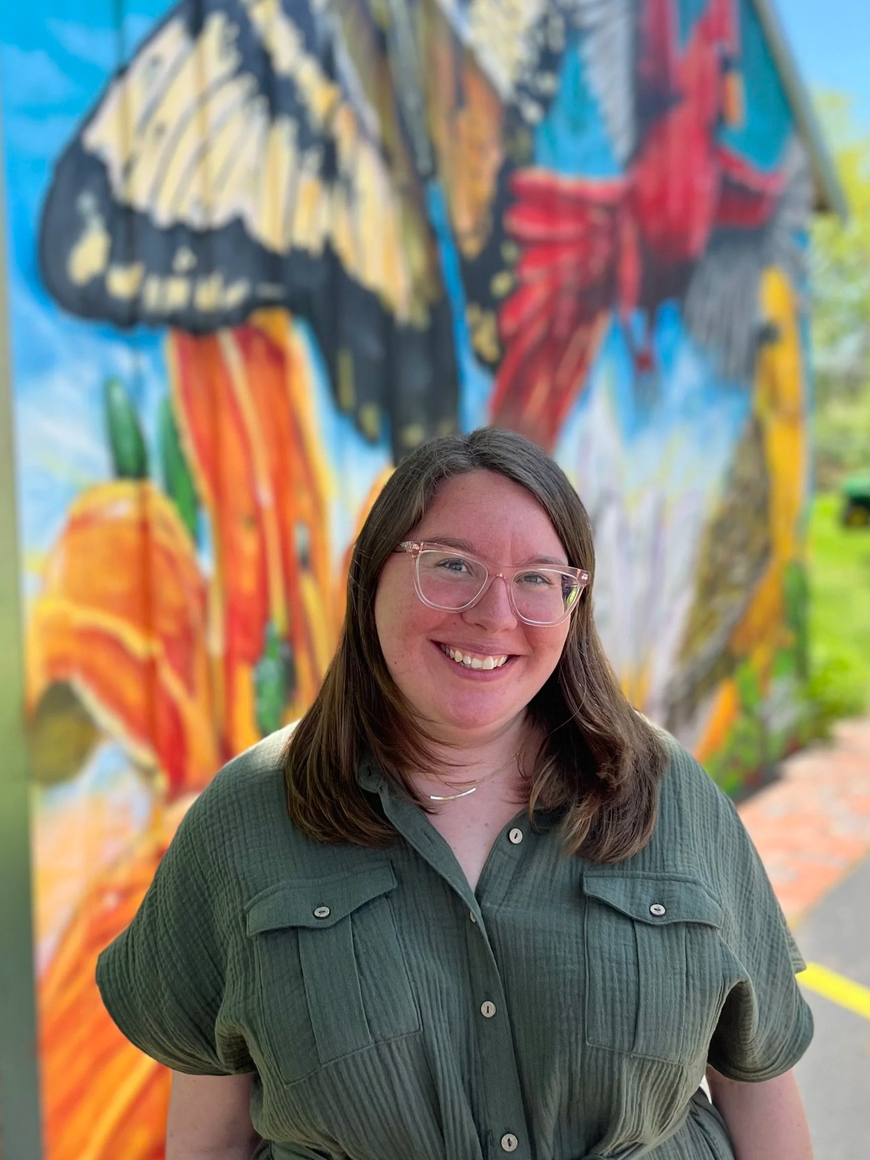 A woman with shoulder-length brown hair, wearing glasses, smiling, standing outdoors in front of a colorful mural featuring abstract shapes in various colors including yellow, red, blue, and black.