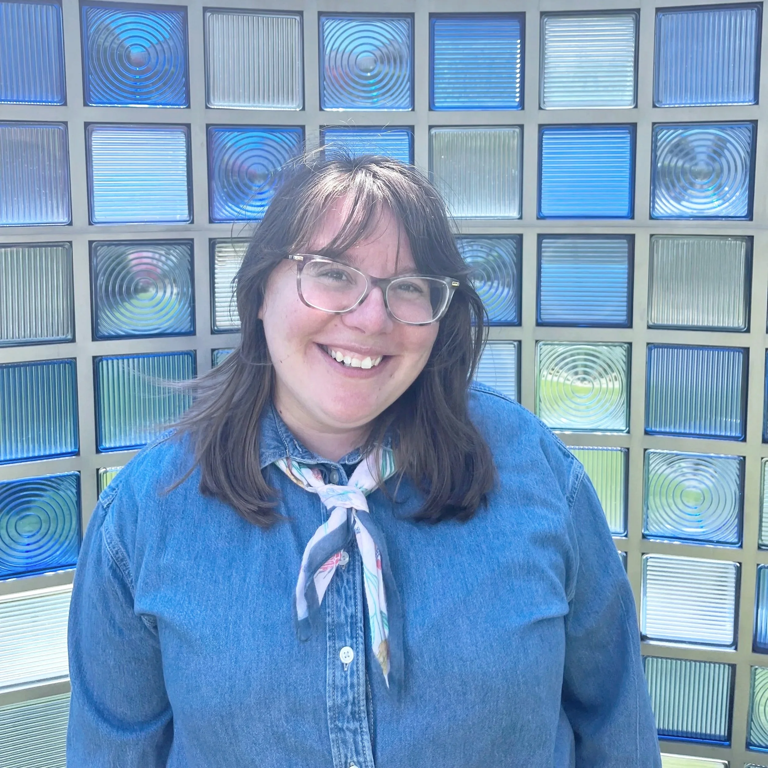 Close-up of a smiling woman wearing glasses, with shoulder-length brown hair, standing outdoors in front of a background made of multiple textured glass blocks with blue and clear colors.