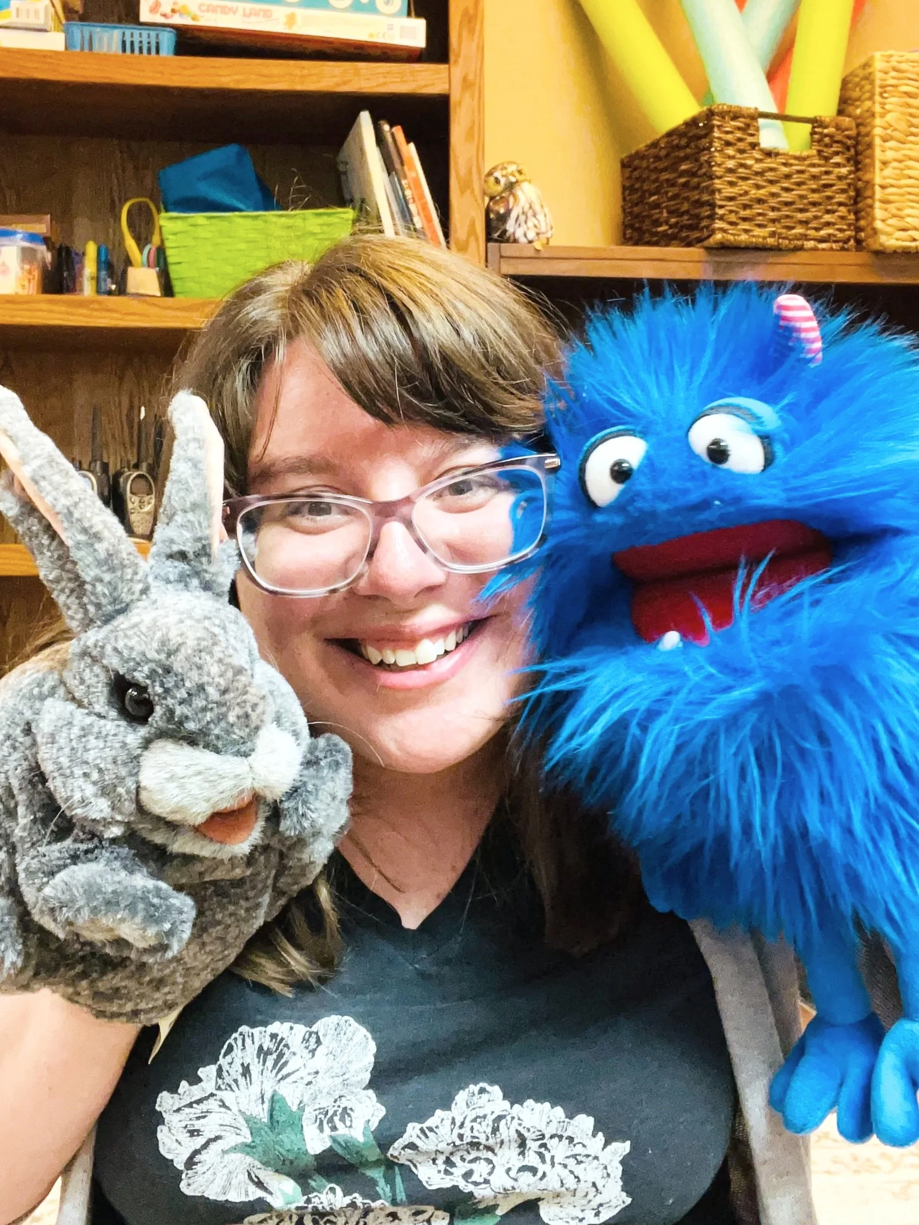 A woman with glasses smiling, holding a gray bunny puppet in one hand and a blue furry puppet with large eyes a in the other.