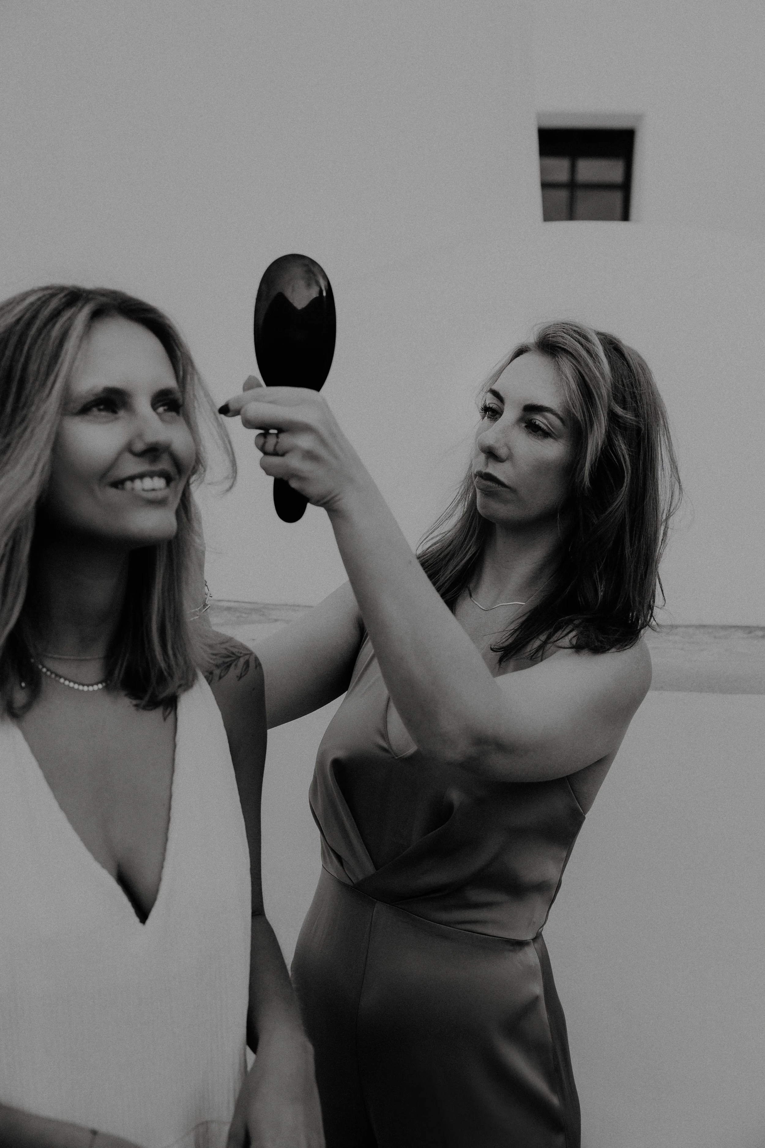 Twee vrouwen, waarvan één kapster die haar de ander haar haar styled. Eén met lange, golvende haar en een sieraden ketting, de andere met stijlvol haar en een jurk, poseren samen terwijl een van hen zich in een kleine handspiegel bekijkt