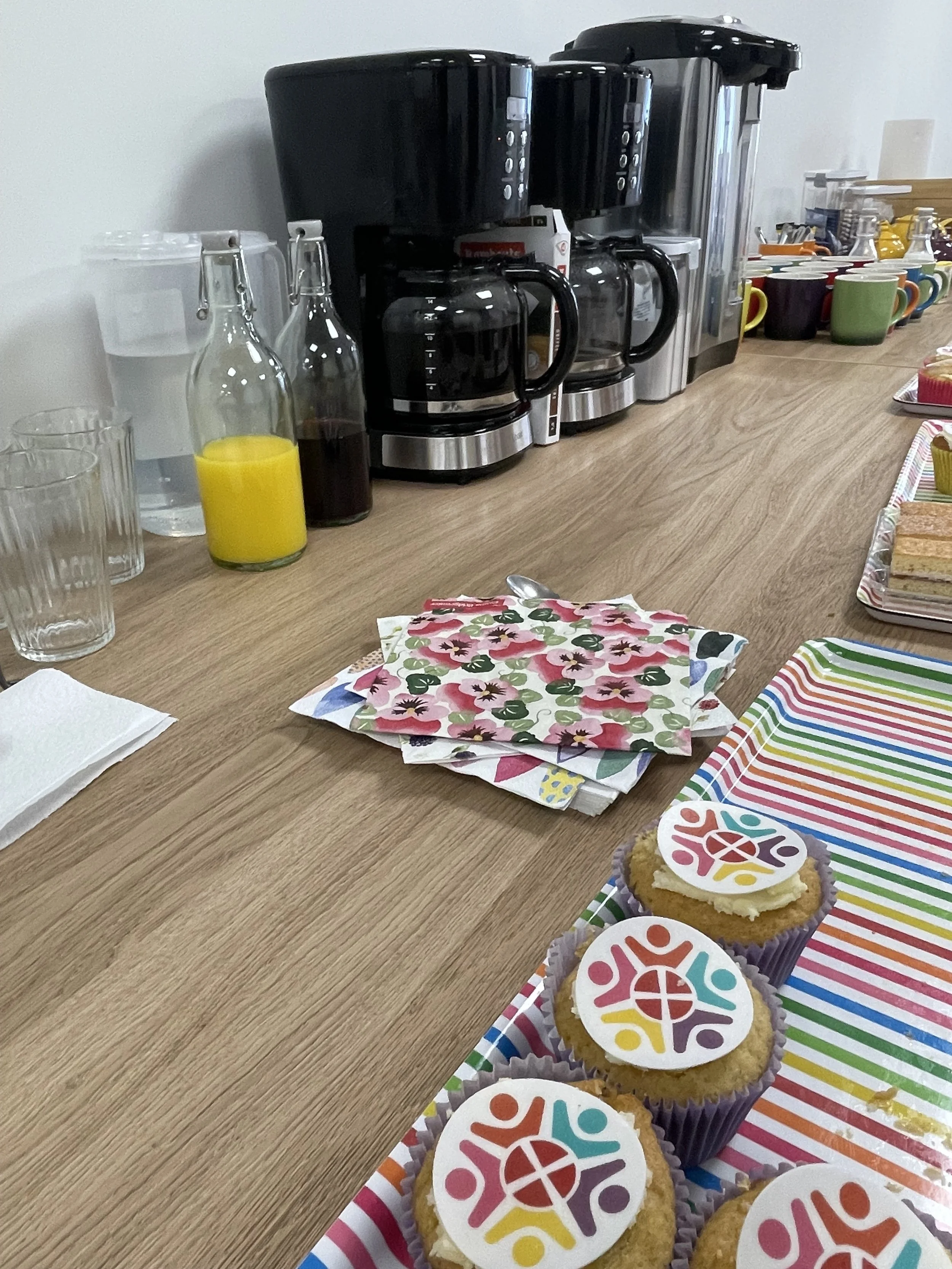 Kitchen counter with three coffee makers, glass containers, colorful mugs, and cupcakes with colorful logos.