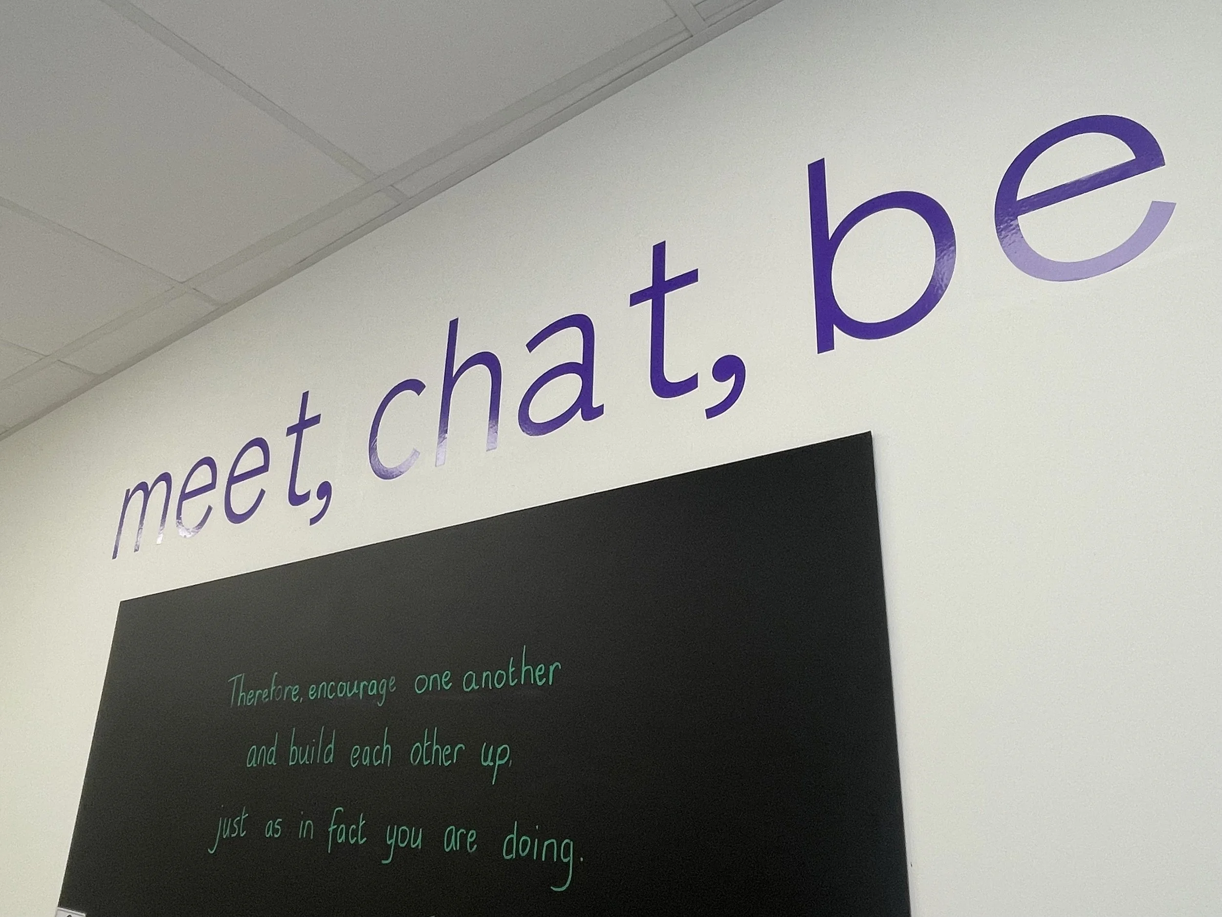 The image shows a wall with motivational quotes written in colorful chalk. The visible part of the quote reads, "meet, chat, be" in purple and partly visible text below it, which is green, says, "Therefore, encourage one another." The background of the wall is white and there is a black chalkboard.