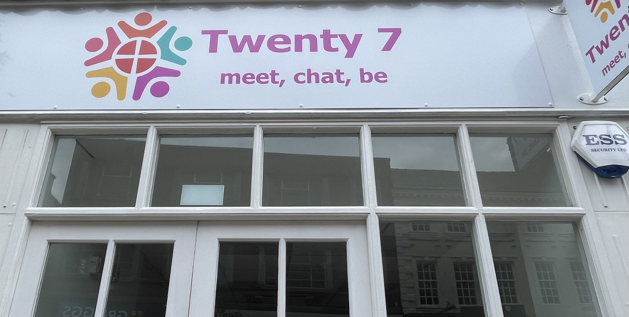 Exterior of a building with a sign reading "Twenty 7 meet, chat, be" and colorful logo, with a security alarm box on the right side.