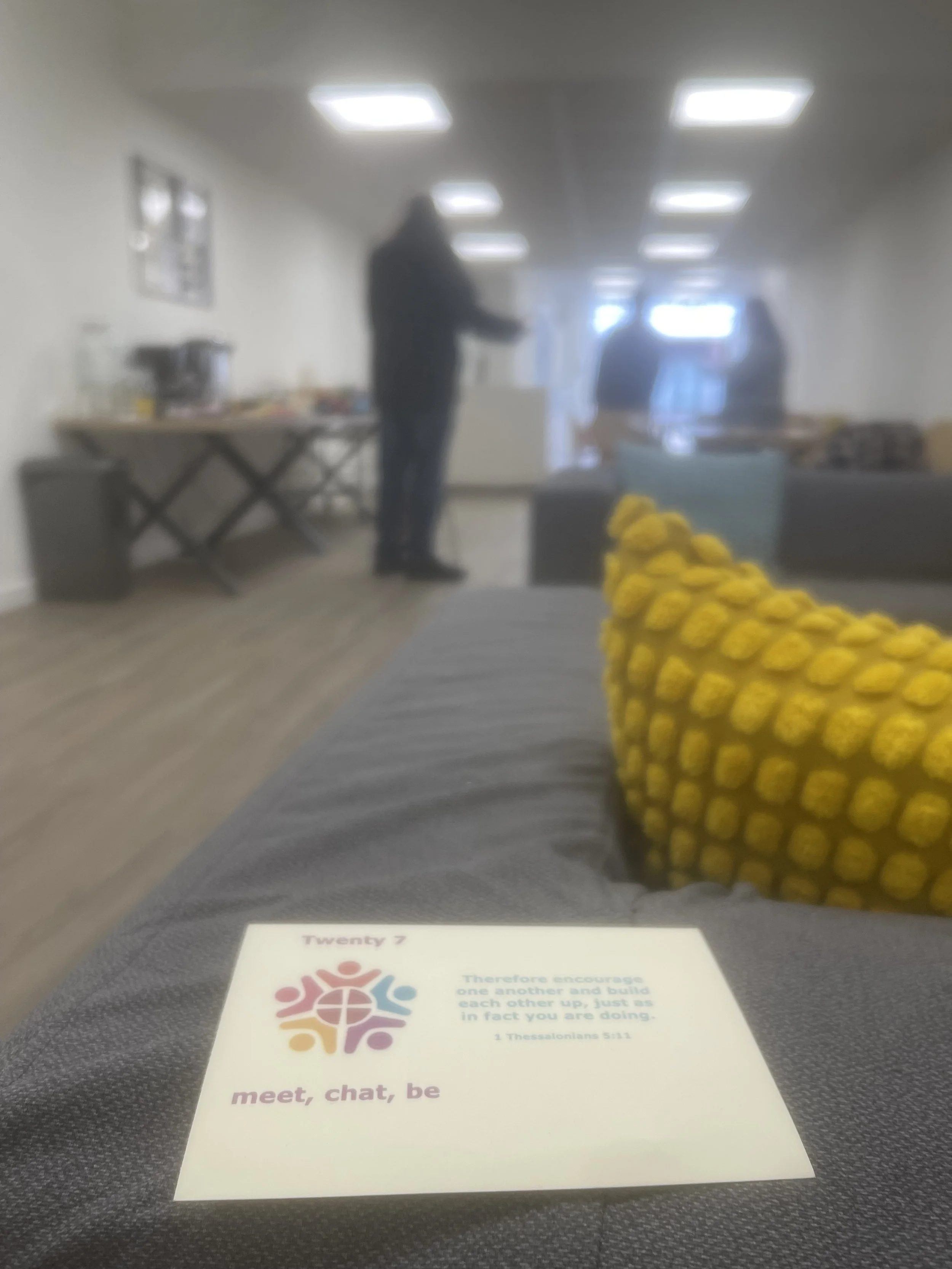 A blurry indoor scene with a person standing and talking, a table with food or items, a couch, some wall hangings, and a sheet of paper with a colorful logo and text on it in the foreground.