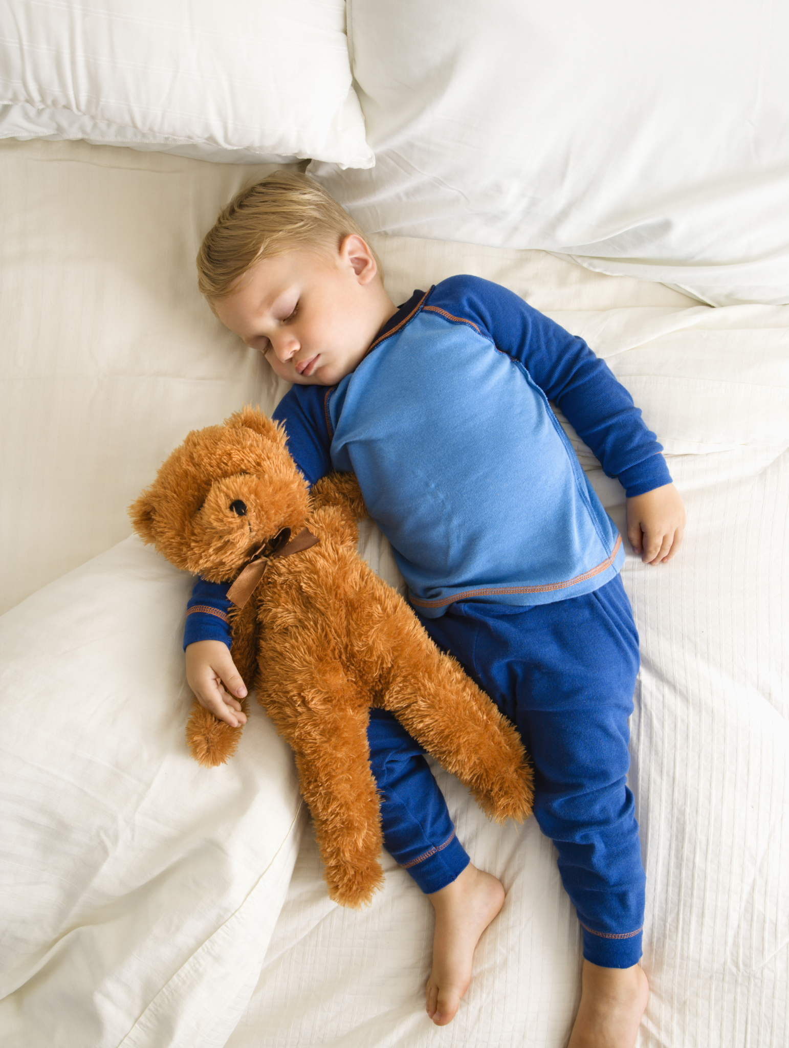 A young boy with blond hair sleeping on a white bed, cuddling a large brown plush teddy bear.