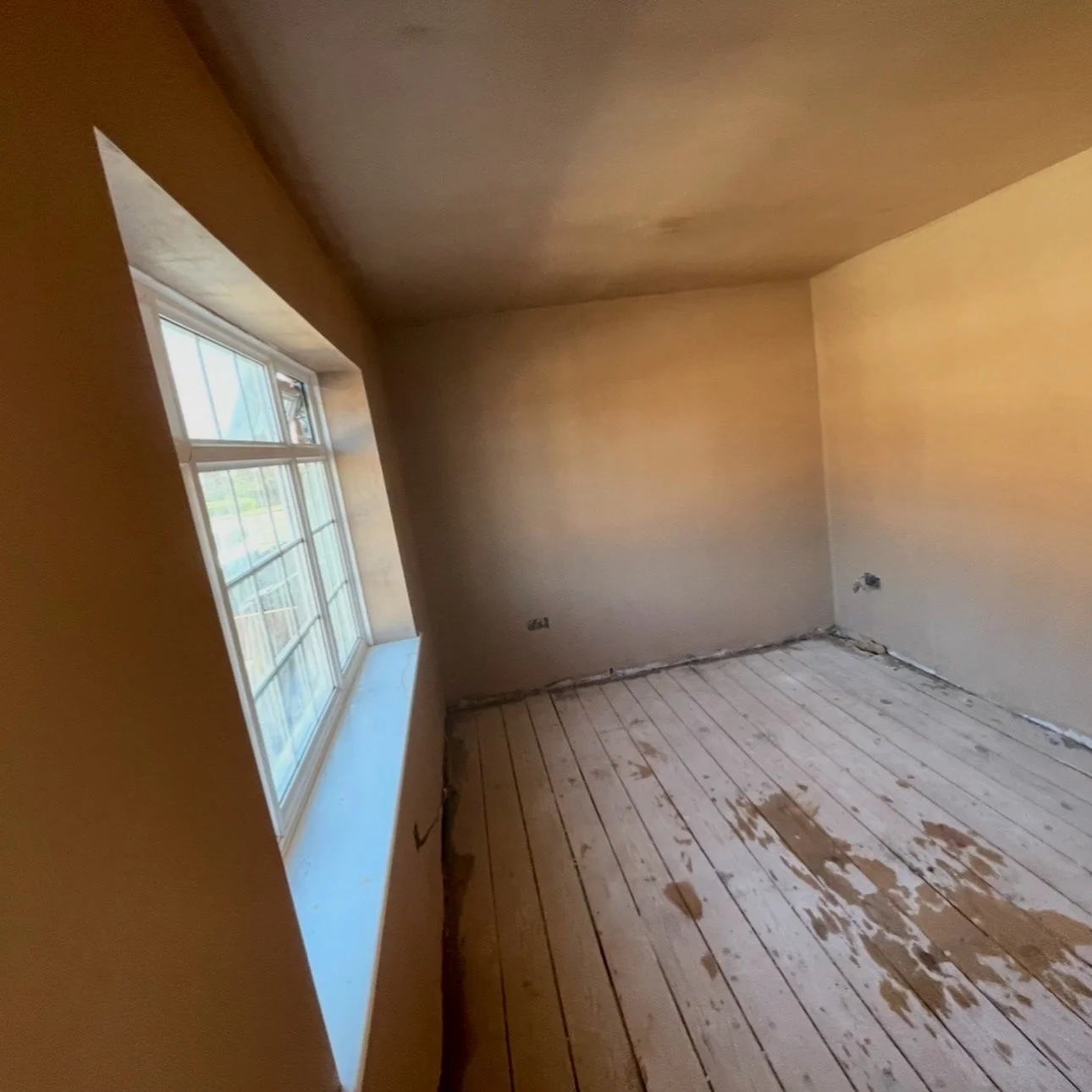 Empty room under construction with wooden floorboards, beige walls, and a window on the left side, allowing natural light inside.