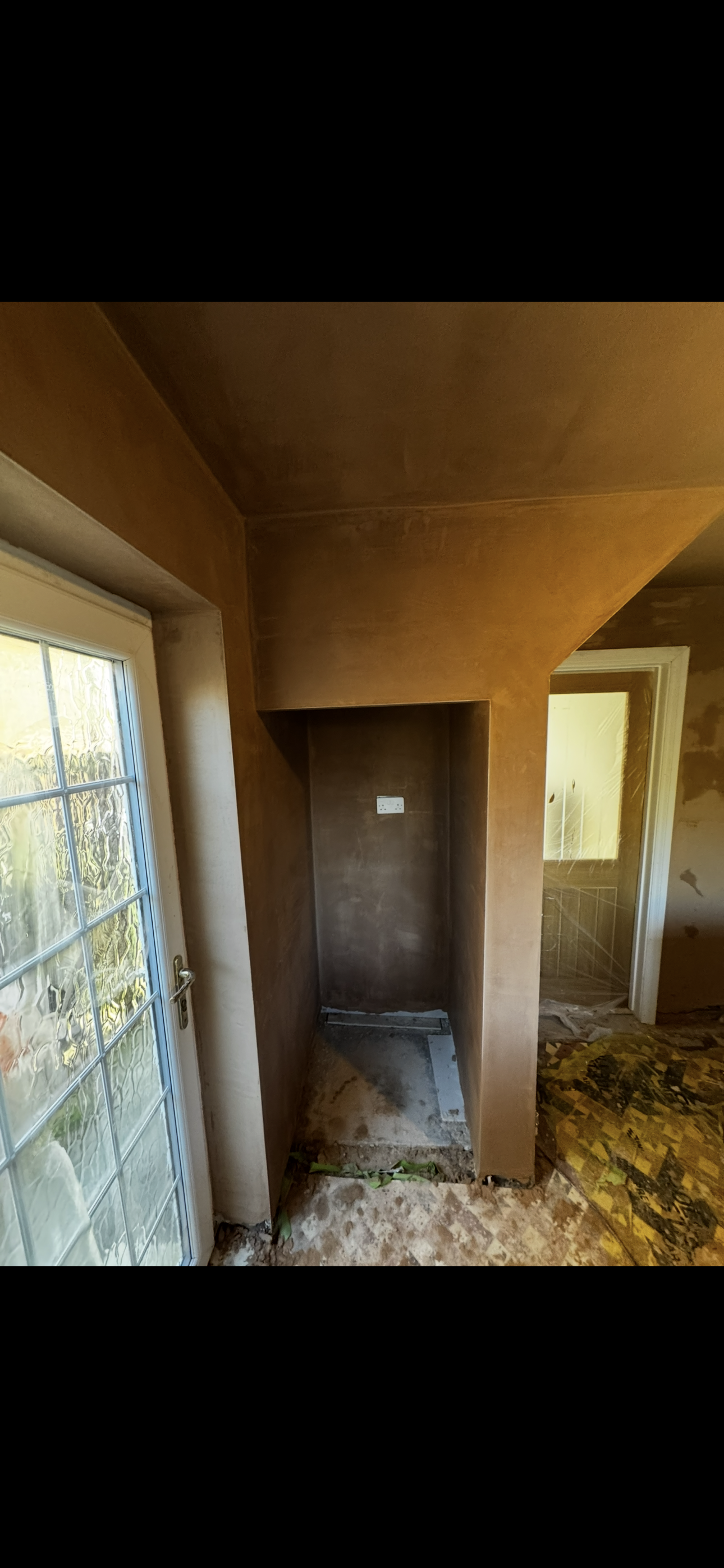 Interior of a room under renovation with brown walls and ceiling, an electrical outlet on the wall, and a window with a door on the left side. The floor is unfinished.