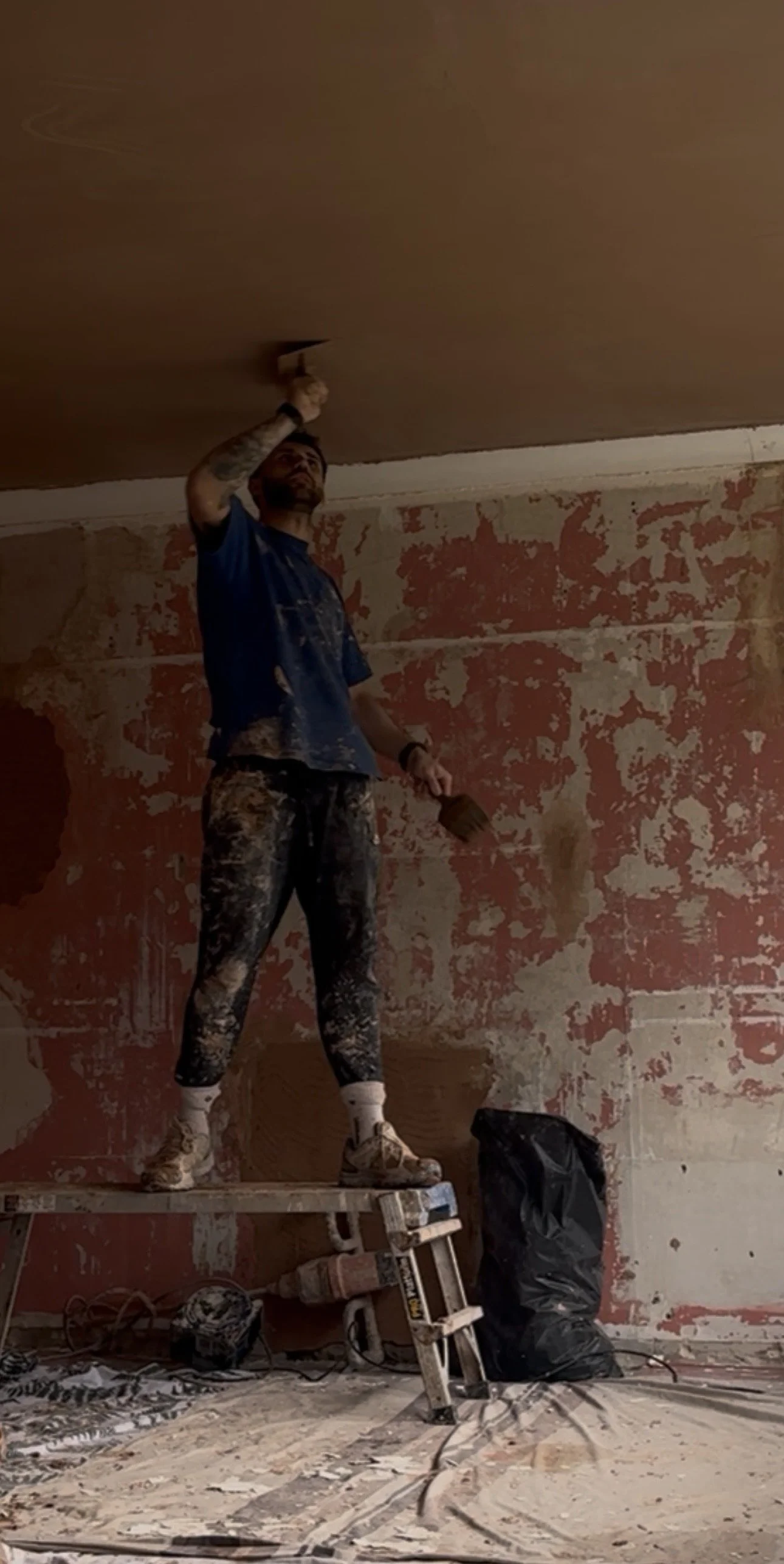 A man standing on a step ladder, painting a ceiling in a room with peeling paint on the walls. He is wearing paint-splattered clothing and holding a paintbrush while reaching up to paint the ceiling.