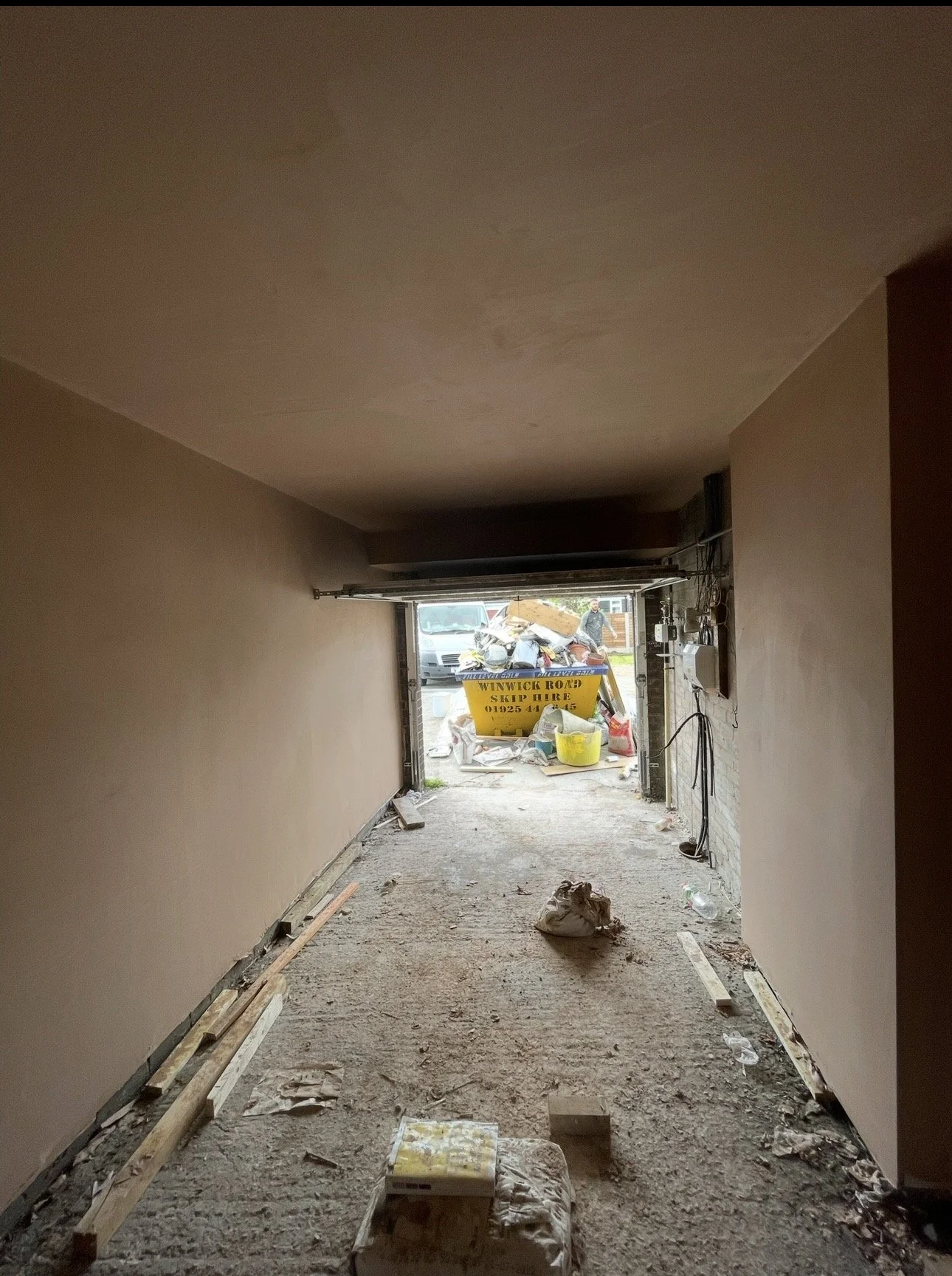 A garage under renovation with drywall on the walls, construction materials on the floor, and a view outside through the partially open garage door, showing a yellow skip and a man walking outside.