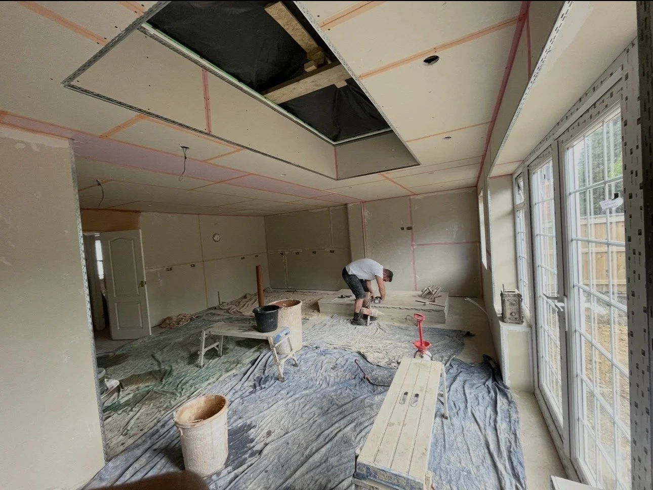 A person working on construction or renovation inside a house. The room has large windows, construction materials, tools, and a protective plastic sheet on the floor.