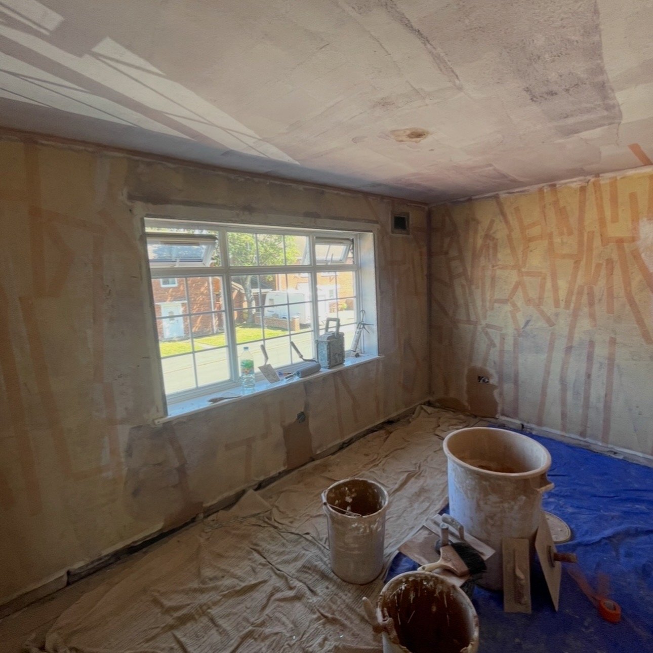 Interior of a house under renovation, with unfinished walls, a large window, and construction supplies including buckets, paintbrushes, and tools on the floor covered with protective sheets.
