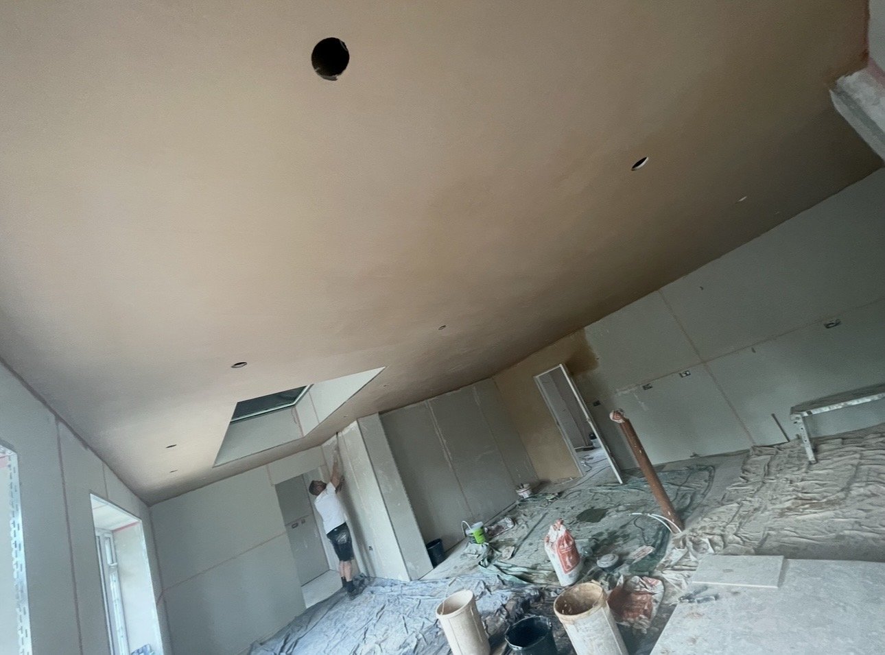 Interior of a room under construction with drywall walls, a partially finished ceiling with holes for lighting, and construction tools and materials scattered on the floor.