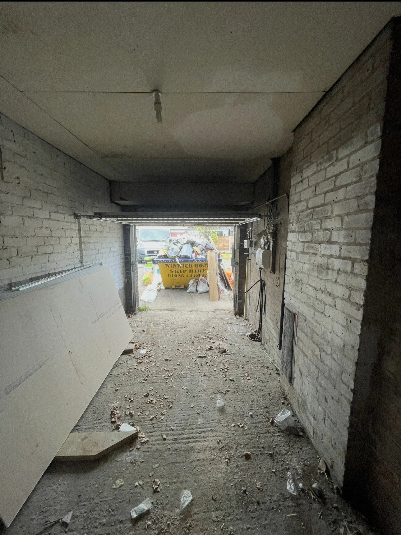 Construction site inside a garage or similar structure, with brick walls, exposed wiring, drywall leaning against the wall, a dirt floor with debris, and an open garage door revealing construction waste and equipment outside.
