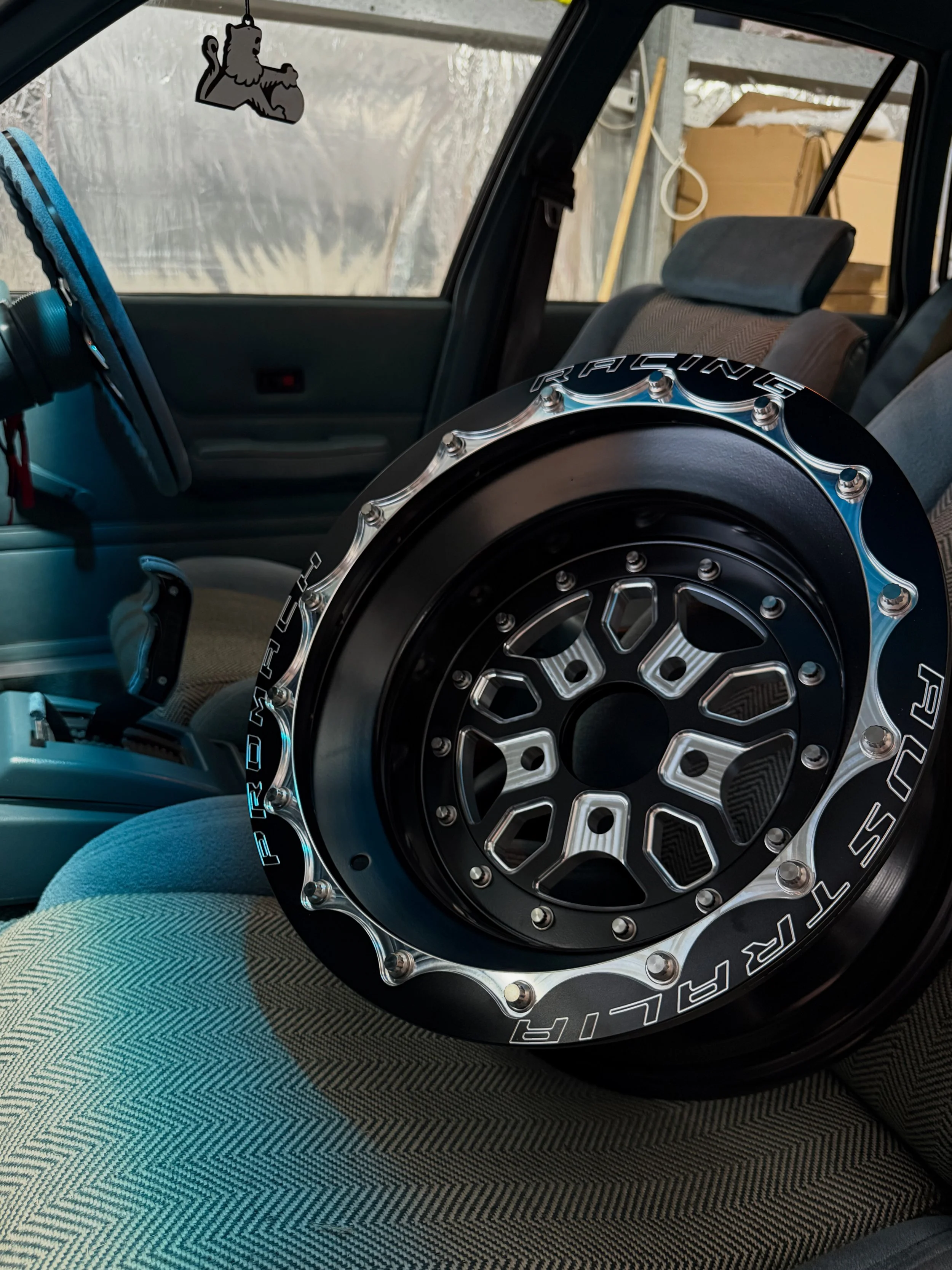 Inside a vehicle, a large black and silver racing wheel is stored on the passenger seat. The vehicle's interior features a gray seat and a black dashboard. A lion-shaped air freshener hangs from the rearview mirror, and the background shows a garage or storage area with cardboard boxes and a wooden structure.