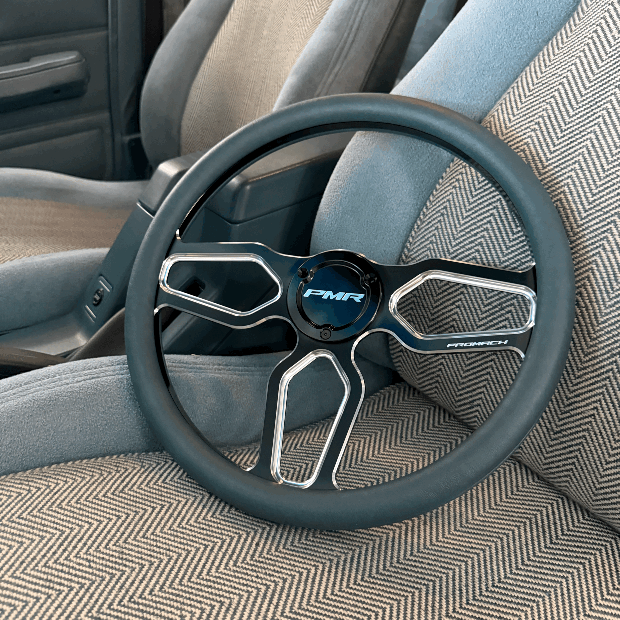 Close-up view of a black steering wheel with 'PMR' and 'Promach' logos, resting on a pink, black, and white herringbone-patterned car seat.