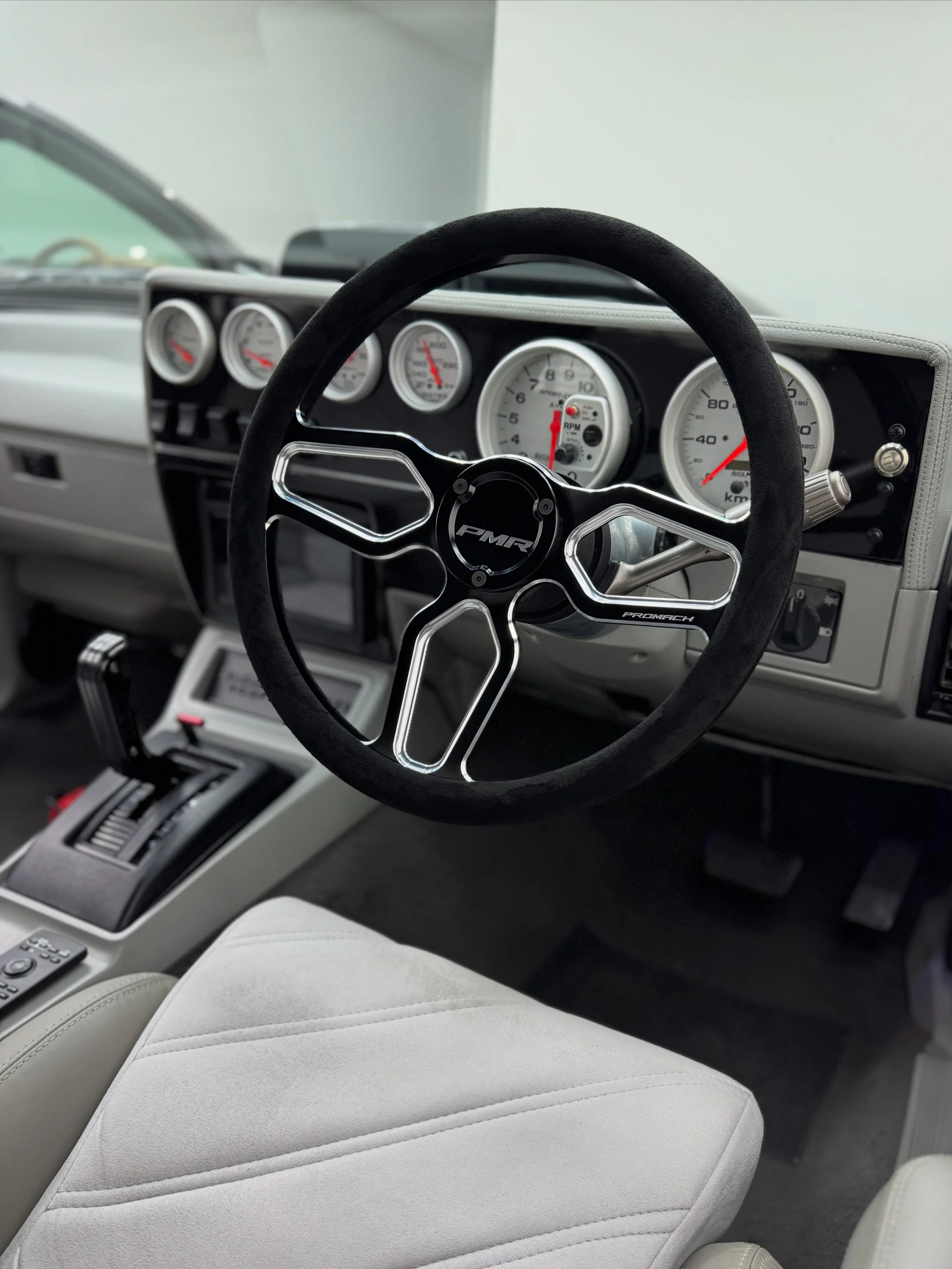 Close-up of a car's interior dashboard featuring a black steering wheel with silver accents, four white gauges with red needles, and a gray center console with various controls, with light beige seats.