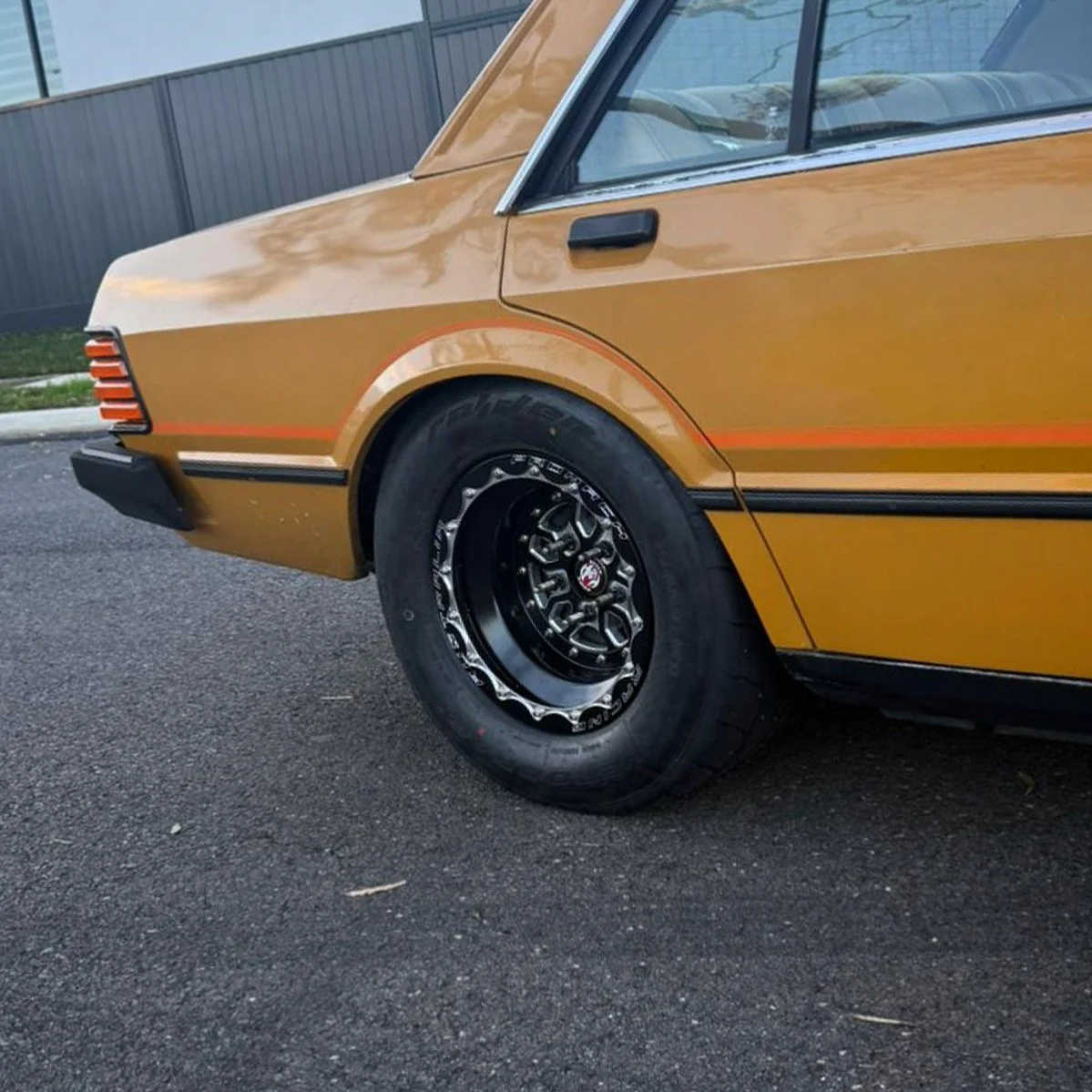 A vintage yellow car, viewed from the rear side, parked on a street. The car has black wheels with chrome accents and a black rear bumper.
