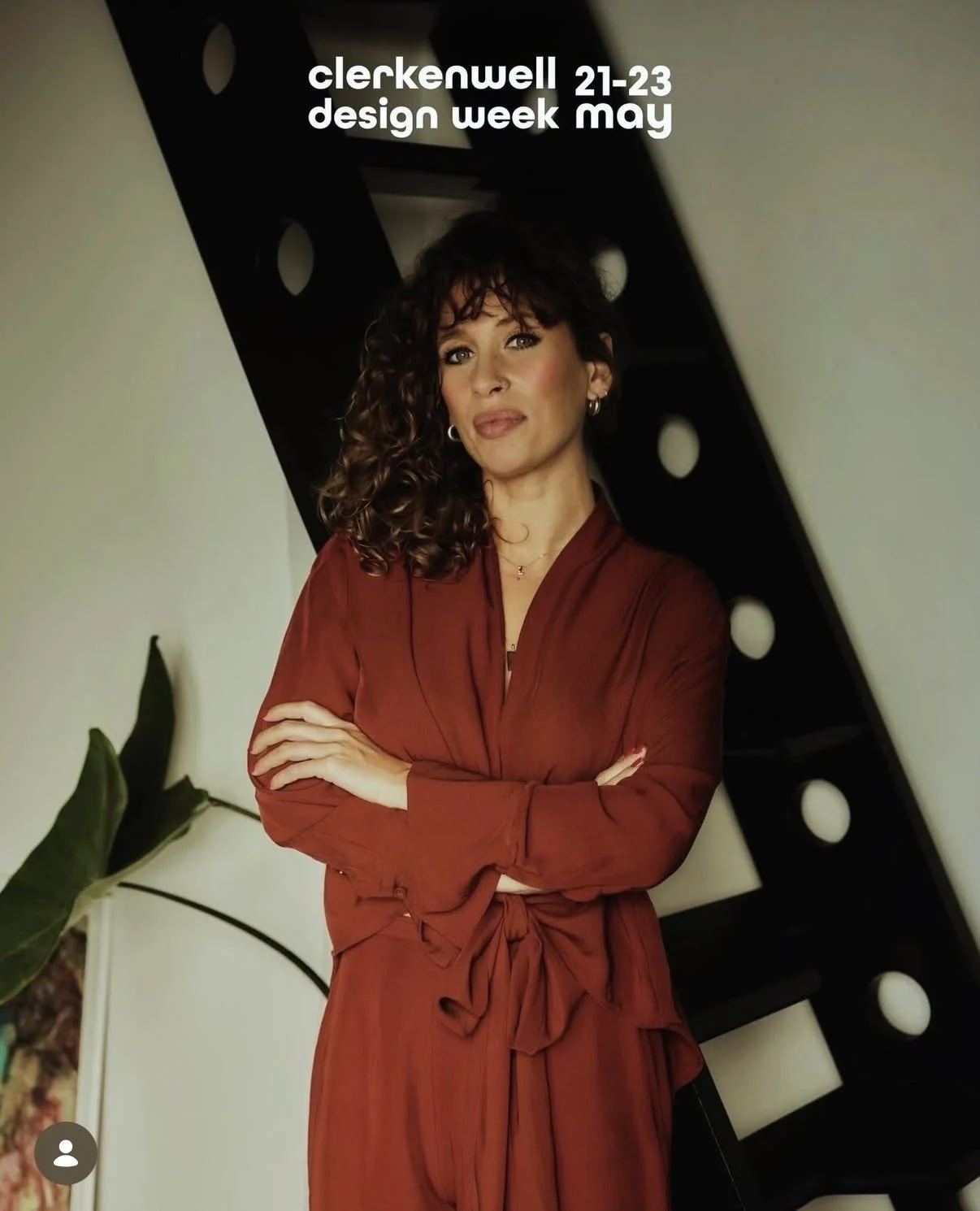 A woman with curly brown hair and hoop earrings standing with arms crossed in front of a black film reel display. She is wearing a rust-colored dress. Text at the top announces Clerkenwell Design Week, May 21-23.