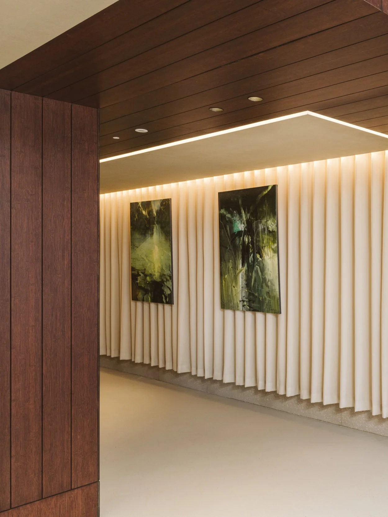 Interior view of a modern gallery or lobby with wooden paneling on the ceiling and a wall covered with cream-colored curtains. Two abstract landscape paintings are hung on the curtains.