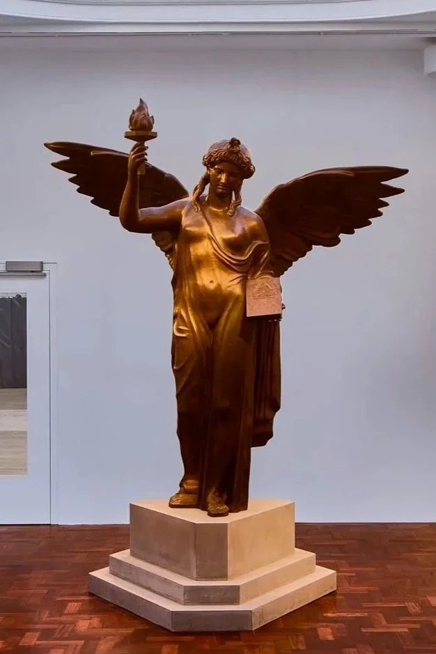 A gold-colored statue of a winged woman, holding a torch in her right hand, standing on a layered base in an indoor setting.